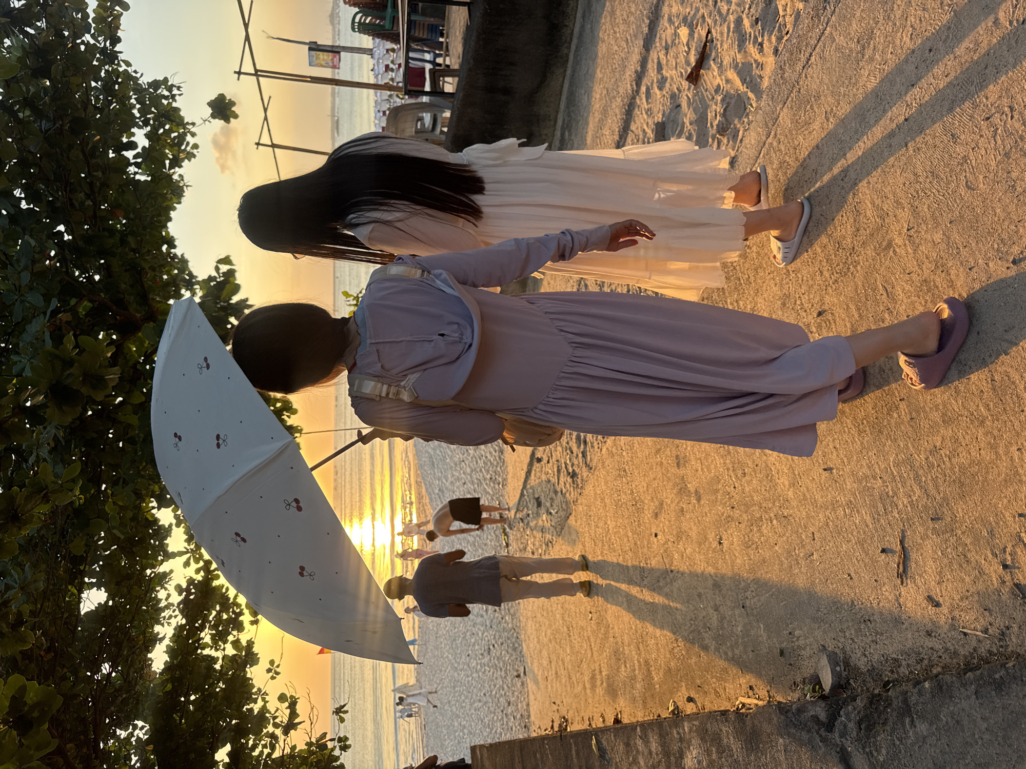 The image depicts a serene sunset scene at a beach in Kuta, Indonesia. Two women, one in a white dress and the other in a purple dress, are walking on a paved path beside a sandy beach. The woman in purple holds a white umbrella adorned with small red cherry patterns. In the background, several people are visible on the beach, some appearing to be engaged in activities like stretching or exercising. The warm, golden hues of the setting sun cast long shadows across the path and sand, creating a tranquil and picturesque atmosphere. The presence of trees and the ocean suggests a coastal location. The overall mood is peaceful and relaxed. No visible text is present in the image.