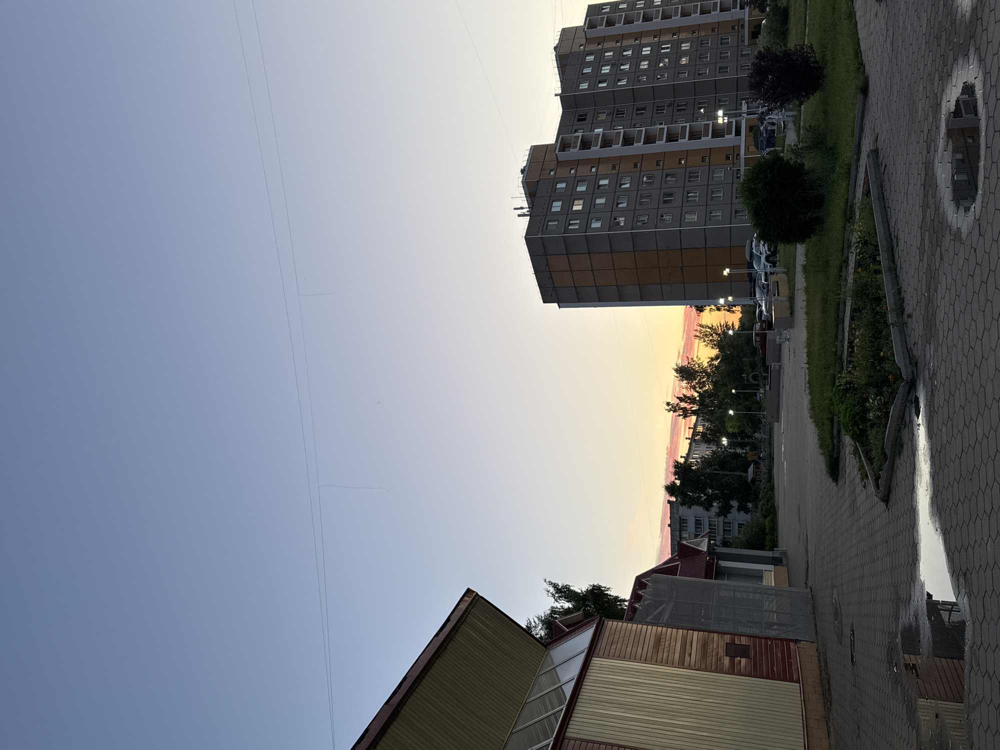 The image captures a scene at dusk in the city of Chaikovsky, Russia. In the foreground, a paved area made of interlocking hexagonal stones features several puddles of water reflecting the sky and surrounding structures. To the left, a building with a red and beige facade and a sloping roof, possibly a sports facility or community center, is partially visible. Further in the background, a tall, grey apartment building with orange accents and numerous windows stands prominently. Between these buildings, trees are scattered, and a parking lot with several cars is visible. The sky is a gradient of pale blue and soft orange hues, indicative of sunset. Streetlights are illuminated in the distance. There are no people or animals visible in the frame. The overall impression is a quiet, residential urban landscape at the end of the day.