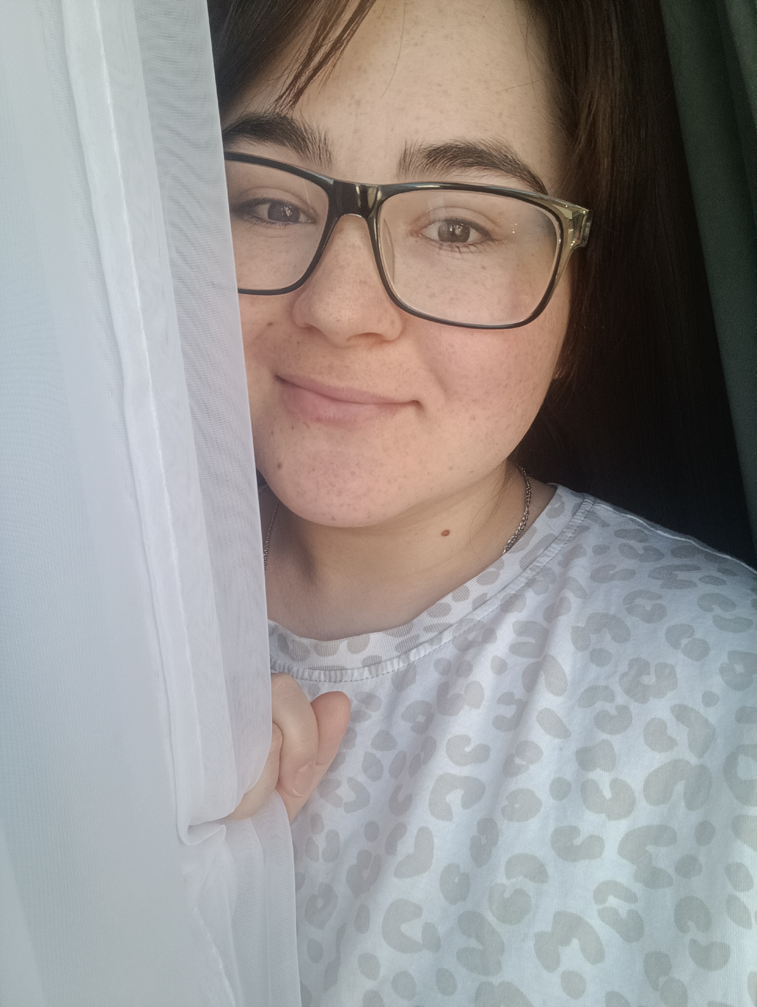 A close-up portrait shows a young woman peeking from behind a sheer white curtain in what appears to be an indoor setting in Noviy, Russia. Her face, adorned with freckles, is partially visible on the right side of the frame. She wears dark-rimmed eyeglasses and looks directly towards the viewer with a gentle, soft smile. Her dark hair is visible around her head, and her eyebrows are dark. Her right hand, holding the curtain, is seen in the lower left portion of the image.
She is wearing a light-colored, possibly white or very light grey, t-shirt with a subtle, lighter grey leopard print pattern. A thin silver chain necklace is visible around her neck, and a small mole is present on her neck below her jawline. The white curtain creates a bright, soft-focused element on the left, suggesting daylight is coming from that direction. A dark, indistinct background is visible behind her on the right. No visible text is present in the image. The overall mood is calm and friendly.