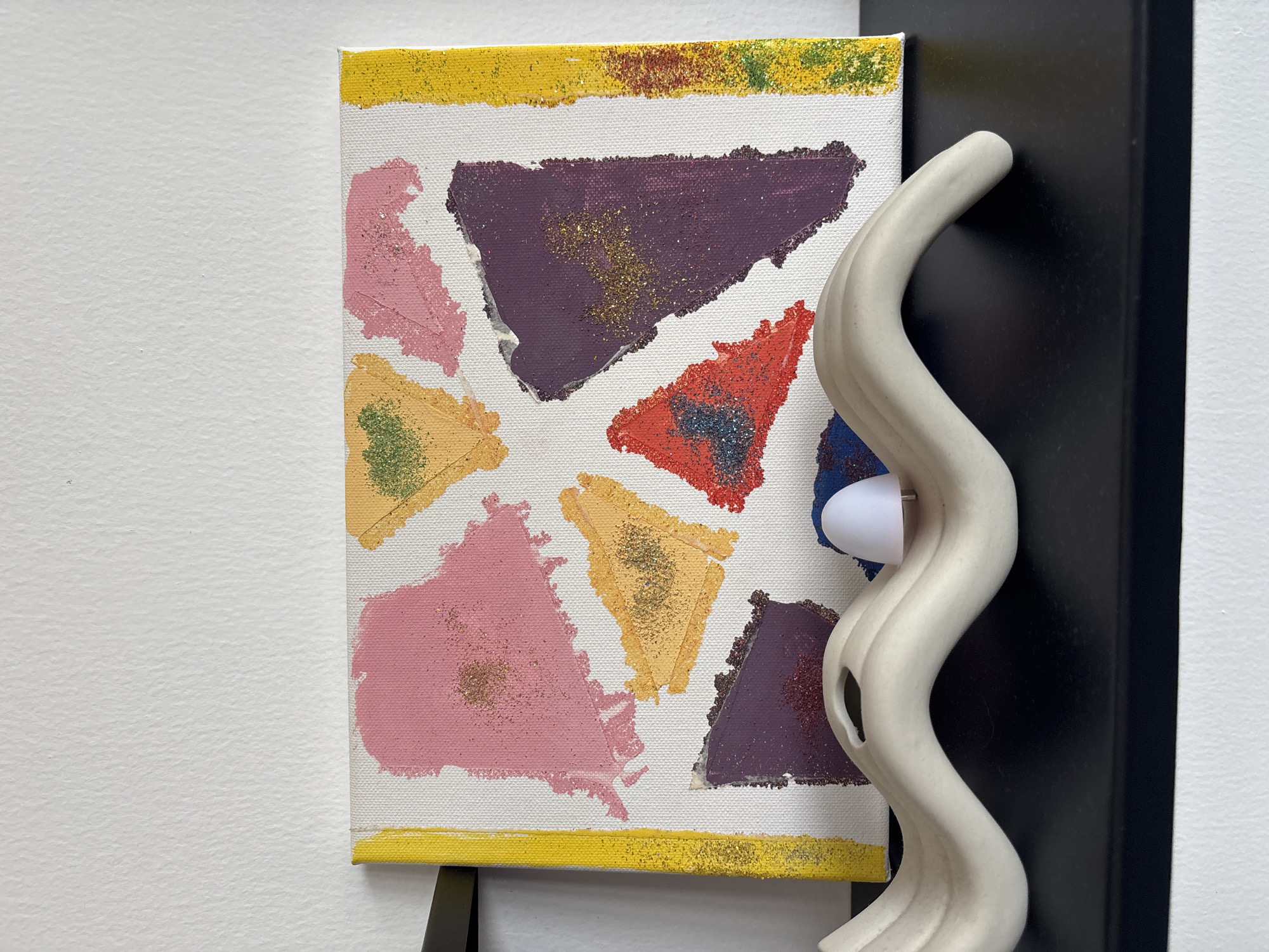 A close-up, eye-level shot shows an abstract painting on a canvas featuring various colored triangles with glitter accents. The painting is bordered by yellow on the left and right sides, with the right border also having a thin stripe of red and green. In front of the painting, on a black shelf, rests a beige, wavy sculpture with a small white light fixture on its upper surface. The background is a white textured wall. The image is taken indoors, likely in a residential setting. No specific location in Dubai, United Arab Emirates, can be determined from the visual information provided.