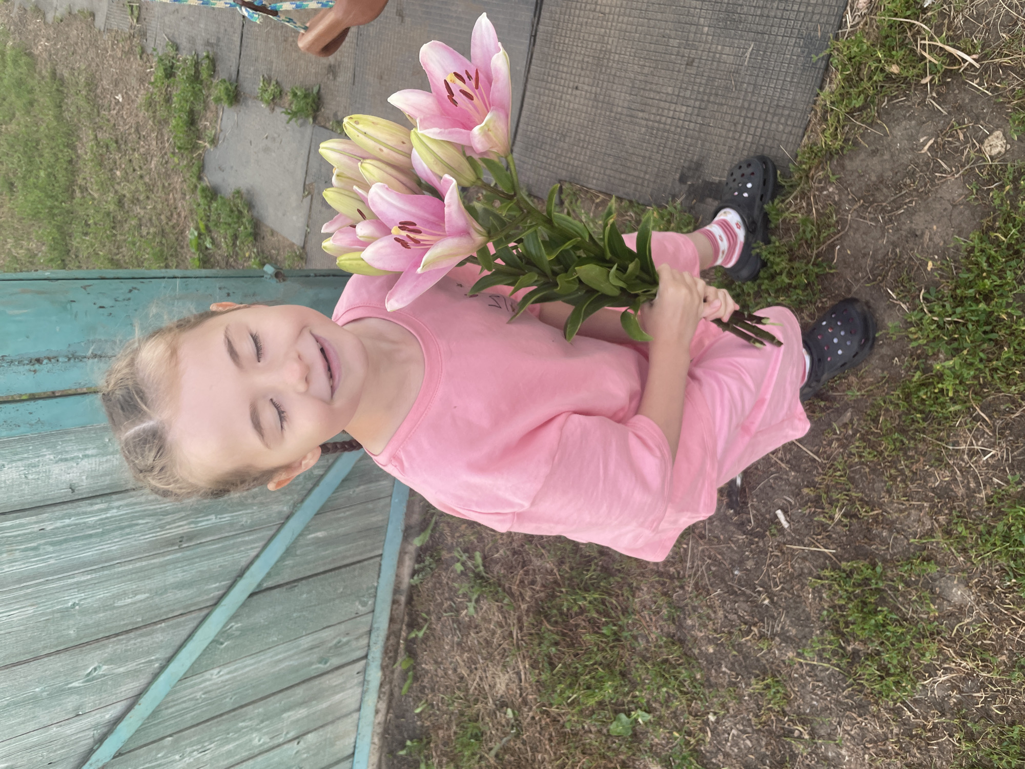 In Nizhnii Bugrysh, Russia, a young girl stands outdoors near a weathered teal wooden fence. She is holding a bouquet of pink lilies. The girl is wearing a pink t-shirt, pink and white striped socks, and black clogs. Her eyes are closed, and she has a joyful expression. The ground is covered with dry grass and dirt. The background features more grass and a grey, textured surface. The lighting suggests it is daytime.