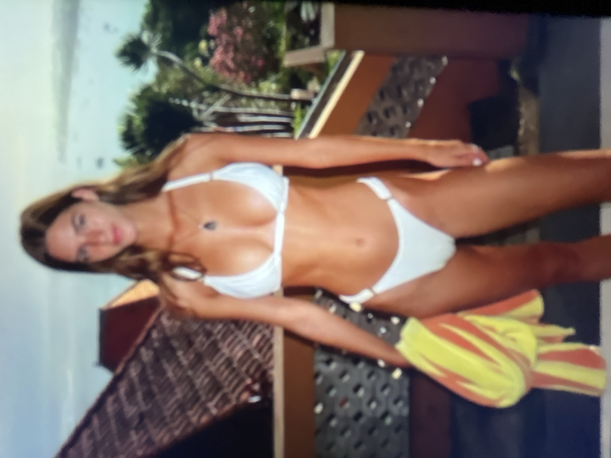 A young woman stands on a balcony in a white bikini, holding a yellow and red striped towel. She has long brown hair and is wearing a necklace. Behind her, a red-tiled roof is visible, along with palm trees and a body of water with boats. The scene appears to be a tropical or coastal location, not the city of Sofia, Bulgaria.