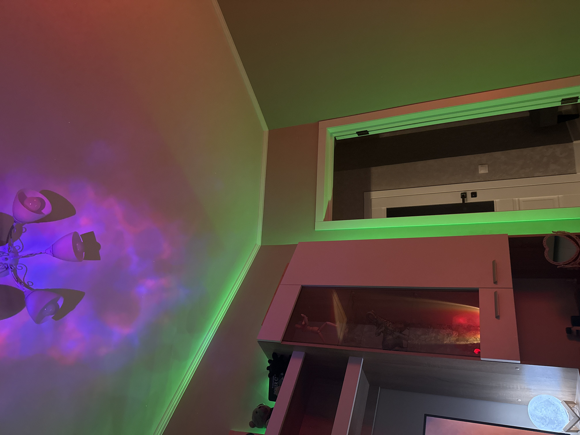 An indoor shot captures a room with a ceiling decorated with colorful light projections and LED strip lighting. A chandelier with multiple light fixtures is visible on the ceiling, casting purple and pink nebula-like patterns. Green LED strips run along the ceiling edge and illuminate a doorway. To the left, a shelf unit holds decorative items, including a teddy bear and a box labeled "Poker". A glass-fronted cabinet displays various objects, one of which appears to be a figurine and another an elephant statue. A moon-shaped lamp sits on a shelf to the far left. A doorway with a white door is visible on the right side of the image. The overall impression is that of a room with ambient, colorful lighting.