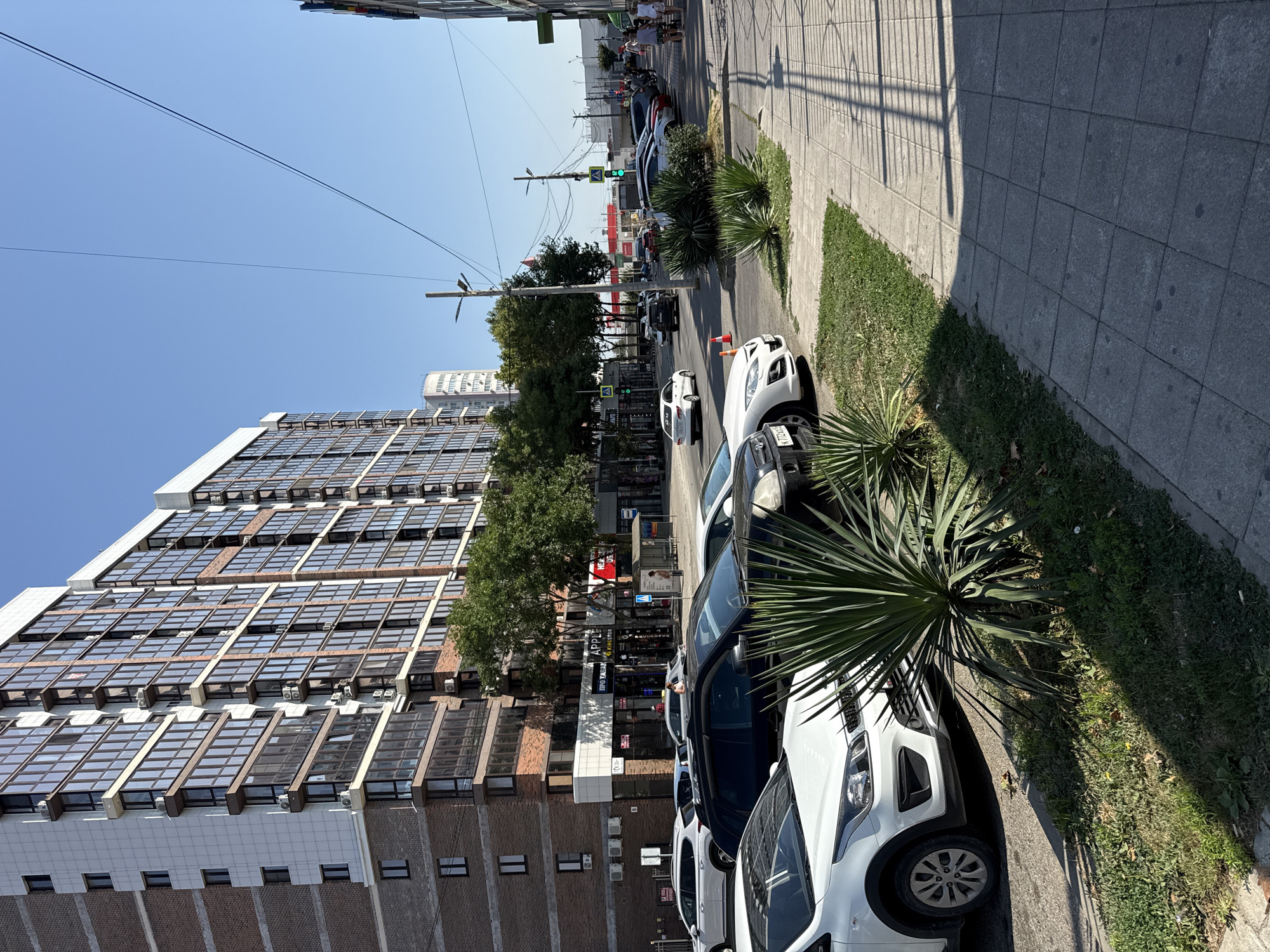 The image depicts a sunny day scene in Anapa, Russia, focusing on a street lined with a modern, multi-story apartment building and commercial establishments. In the foreground, several cars are parked along the street and on the sidewalk. A white Kia Rio is parked prominently on the left, with a dark SUV and other vehicles partially visible. A patch of green grass with spiky plants separates the sidewalk from the street. The apartment building is characterized by its brick facade and numerous glass-fronted balconies. The lower levels of the building feature shops, one with signage for "APPLE KINGSTON" and another with Russian text that appears to be an advertisement. Further down the street, traffic lights are visible, and more cars are parked or driving. Several pedestrians are walking on the sidewalk. The sky is clear blue, indicating fair weather.