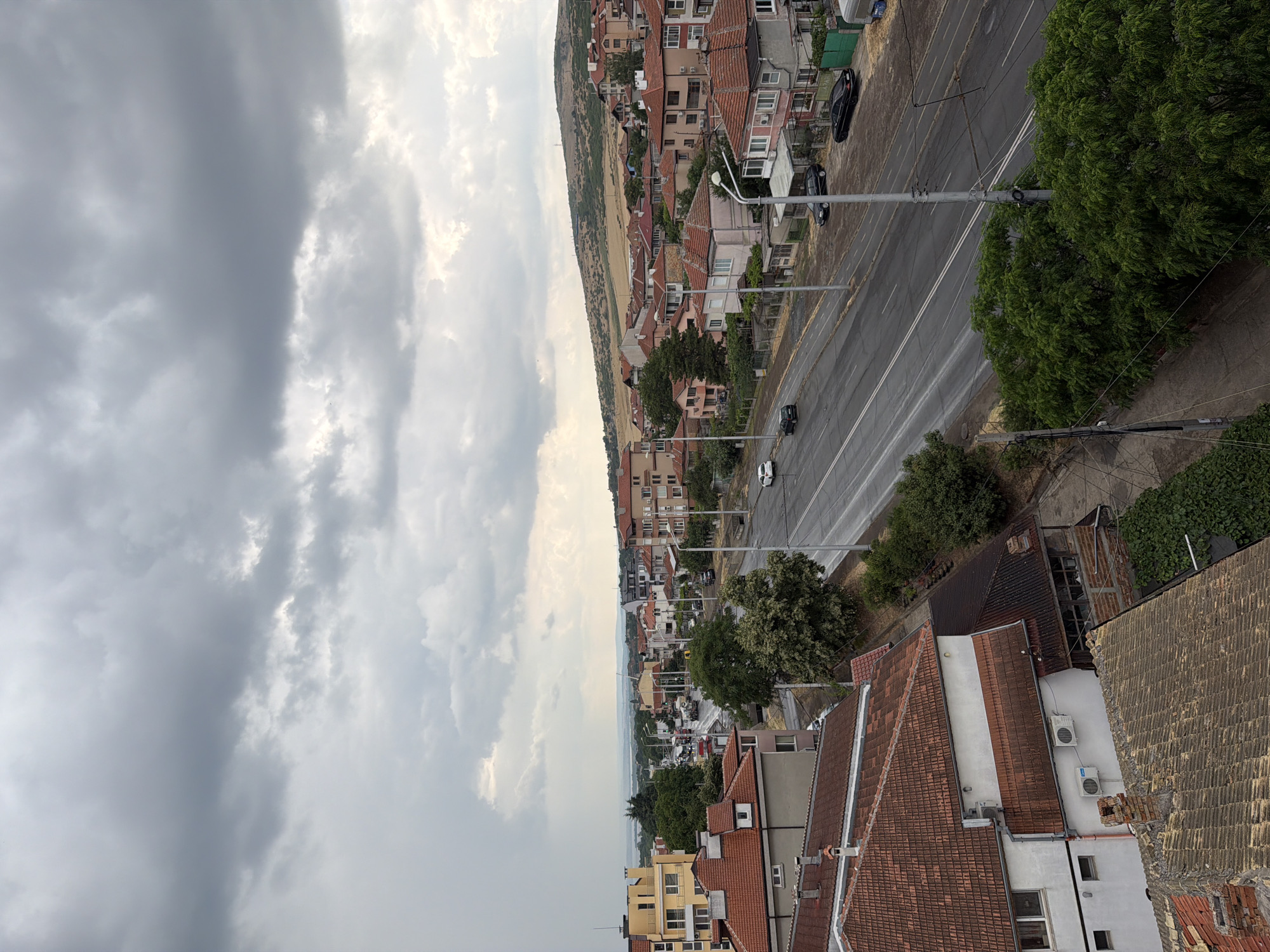 The image depicts a panoramic view of a residential area in Burgas, Bulgaria, under an overcast sky. The scene is dominated by a street with two lanes of traffic, flanked by buildings with red-tiled roofs. Several cars are visible on the street, and some parked on the side. The buildings are a mix of styles, with some appearing older and others more modern. Lush green trees are interspersed throughout the neighborhood, adding a natural element to the urban landscape. In the background, a hill with sparse vegetation rises, and beyond that, a body of water, likely the Black Sea, is faintly visible. The overall impression is of a quiet, slightly somber day in a coastal Bulgarian city.