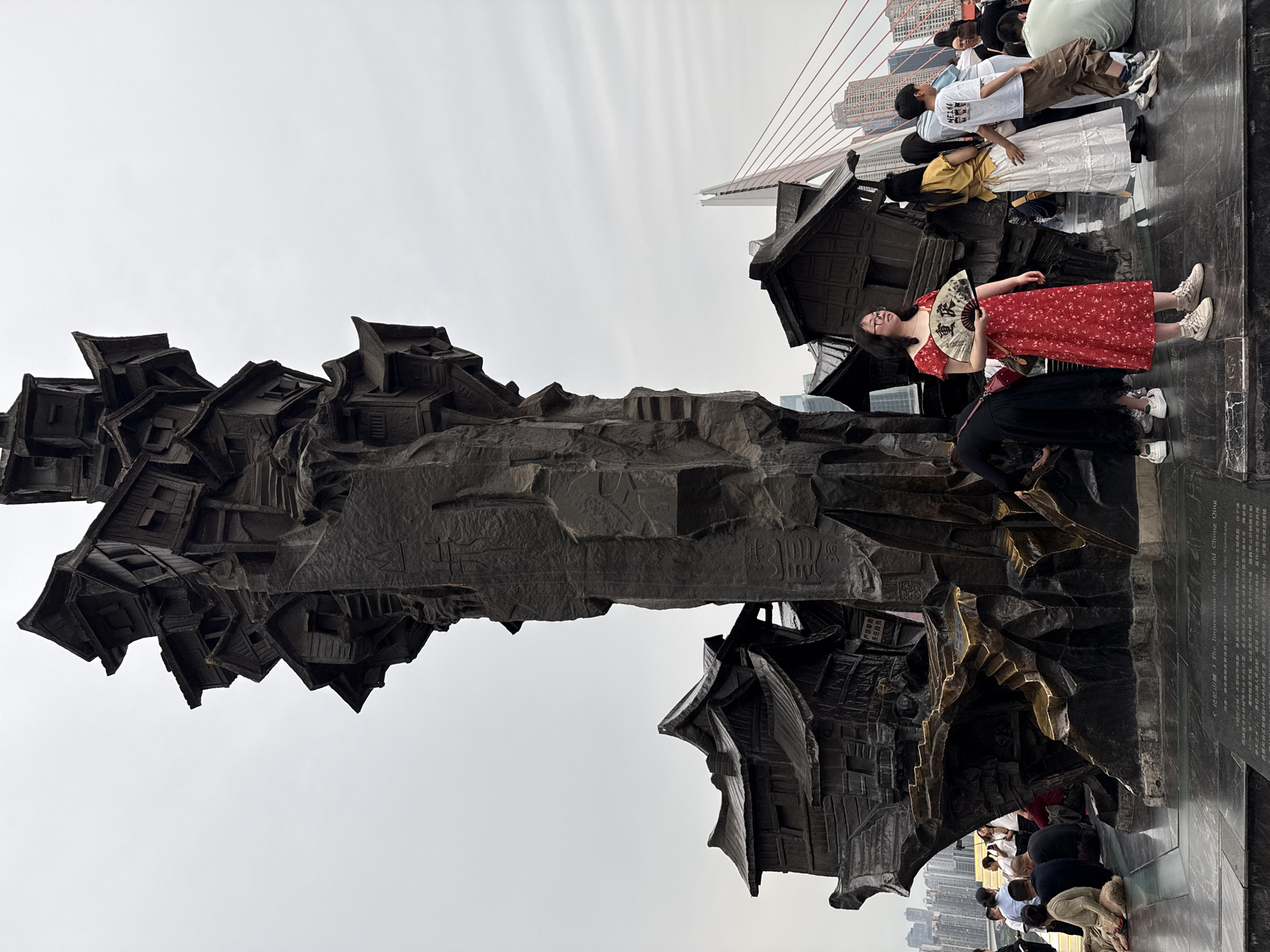 The image shows a large, dark-colored sculpture resembling a cluster of traditional Chinese houses, possibly made of bronze or a similar metal, located in Yuzhong District, China. In the background is a large suspension bridge with red cables, indicating the urban environment. A group of people are standing and posing around the sculpture. One woman in a red dress and white sneakers is holding a fan with Chinese characters on it. Other people are gathered on the ground near the base of the sculpture. The sky is overcast, suggesting a cloudy day. There is signage nearby, possibly with information about the sculpture and its creator, Guo Xuanchang, and it is called "The Impression of the old Chong Qing."