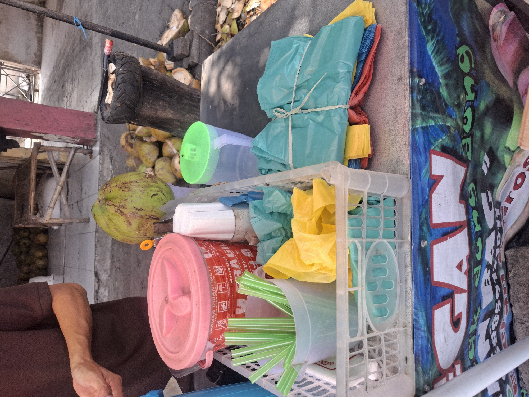 The image depicts an outdoor scene in the city of Karanggeneng, Indonesia, likely a street-side vendor stall. In the foreground, a table is laden with items for sale. A white plastic basket holds numerous green plastic straws. Next to it are several colorful plastic bags, predominantly teal, yellow, red, and blue, neatly tied together with white string. Behind these bags, a clear plastic jug with a green lid is visible. A large pink lidded container, possibly for storage or ingredients, sits further back. A white paper cup is also present.

In the background, a person wearing a brown t-shirt is partially visible on the left, with their arm extended, suggesting they are the vendor. A large, green coconut is prominently displayed. Scattered around the base of a rough wooden chopping post, which is topped with a tire and used for preparing coconuts, are numerous cut coconut shells and more whole coconuts. The ground appears to be paved, with some debris. In the far background, a wooden bench and a red-painted pillar are visible, along with some structures that indicate an urban or semi-urban environment. The overall impression is of a vibrant, active marketplace setting.