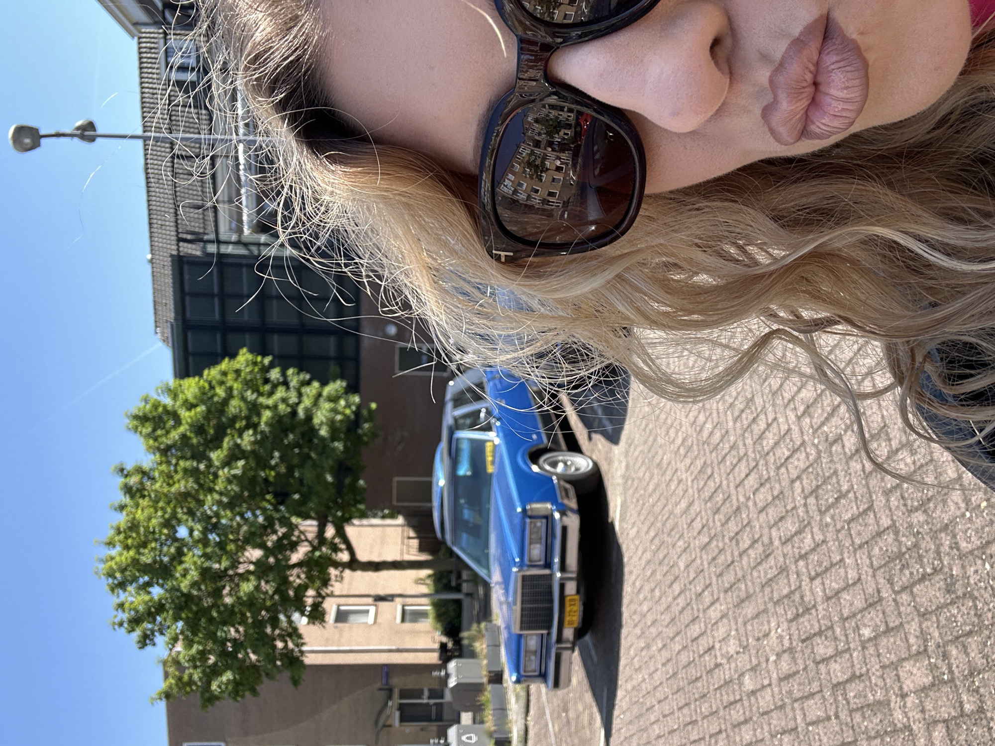 A partially-visible blonde woman with sunglasses is in the foreground of an outdoor scene. Behind her is a blue vintage car parked on a brick-paved street. A tree is to the left of the car, and a building is behind both. The license plate of the car indicates it is in the Netherlands, specifically Amsterdam. A street lamp is visible in the background. The overall impression is that the woman is taking a selfie with the vintage car in Amsterdam.