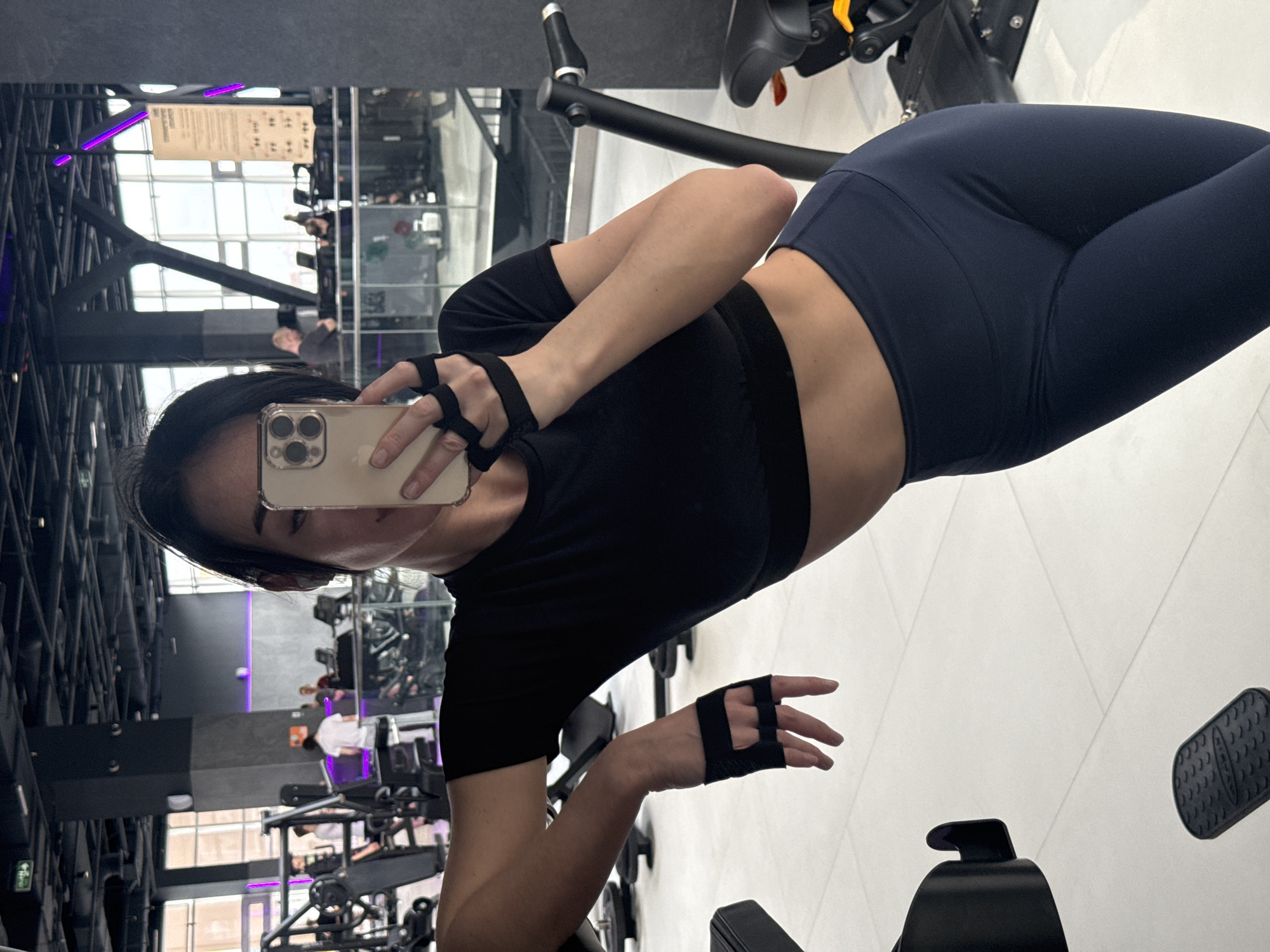 A person, appearing to be a young woman, is captured in a mirror selfie within a modern gym environment. She is positioned slightly to the right of the frame, holding an iPhone with a triple-lens camera system to capture her reflection. Her face is partially visible behind the phone, with dark hair framing her features.
She is dressed in fitness attire, including a black short-sleeved crop top that reveals her midriff, and form-fitting dark blue leggings. Black fingerless gym gloves are worn on both hands.
The background, largely composed of reflections in expansive mirrors, reveals a spacious gym interior in Krasnoyarsk, Russia. Various pieces of gym equipment, such as exercise bikes, ellipticals, and weight machines, are visible. Other gym-goers can be seen in the blurred reflections, some engaging with equipment. The gym features a high ceiling with visible structural beams and integrated lighting, including distinct purple LED strip lights. A large window or glass wall allows for reflections of outdoor buildings. On a reflected sign on a distant wall, blurred text mentions "HOURS" and "CLASS," likely indicating gym schedules or information. The flooring is light-colored, possibly white tiles. The image is a self-portrait taken by the individual, likely during or after a workout session.