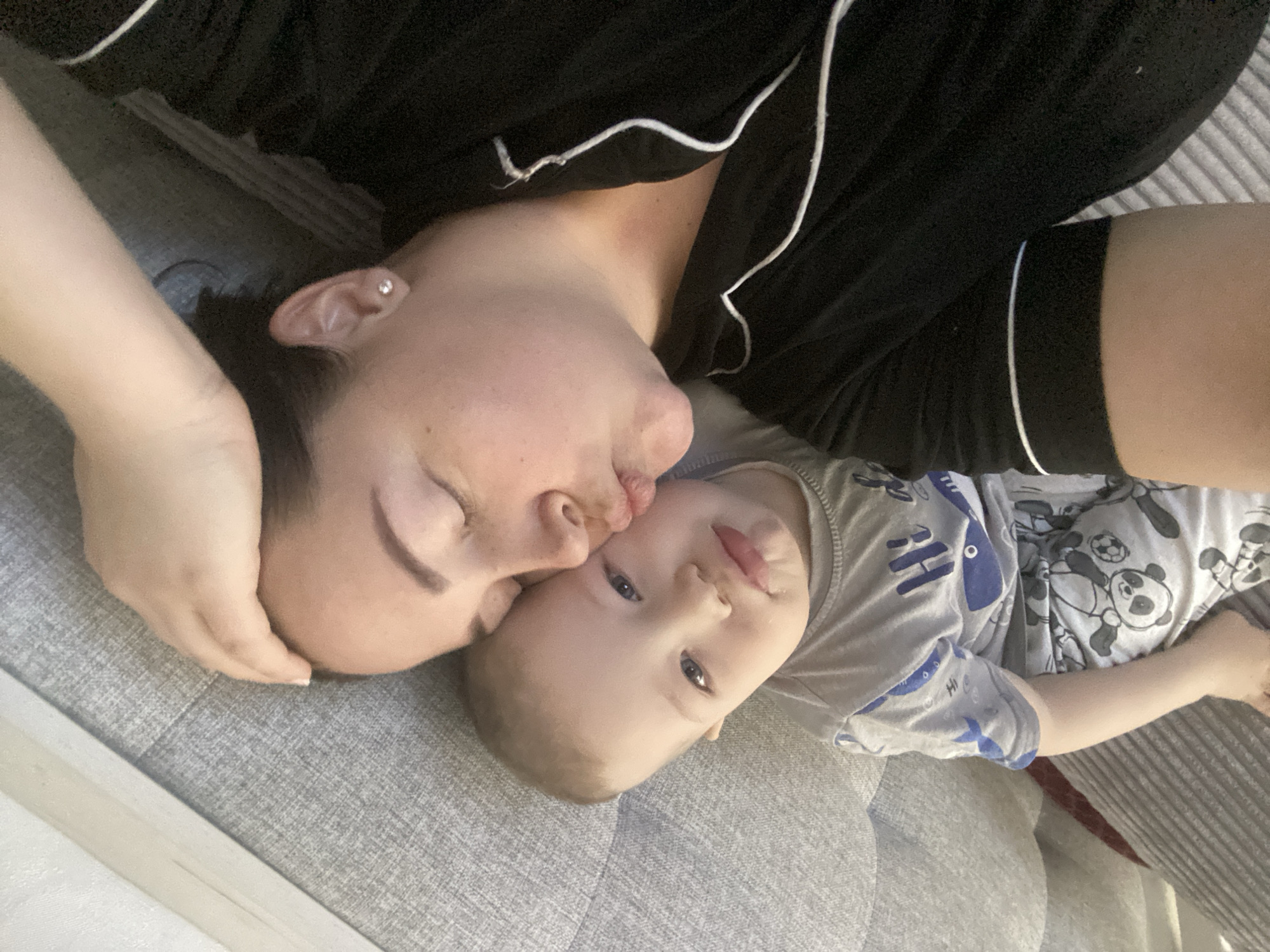 An intimate portrait captures a woman and a young child lying down together, likely on a grey upholstered sofa or bed in Sochi, Russia. The image is taken from an overhead perspective, with the subjects oriented diagonally across the frame.
The woman, with dark hair and a small stud earring visible in her left ear, is wearing a black top with white piping. Her eyes are closed, and her face is pressed against the child's cheek in an apparent kiss or loving nuzzle. Her right hand is gently placed on the back of the child's head.
The child, a toddler with light hair and bright blue eyes, is looking directly at the camera with a relaxed expression. Their mouth is slightly open, revealing a hint of their tongue. The child is wearing a grey t-shirt with blue designs, including the word "HI" and what appears to be cartoon pandas or other animals on the fabric.
The overall mood of the image is one of tenderness and affection, capturing a close bond between the two individuals. The lighting suggests an indoor setting, and no specific time of day is indicated.
