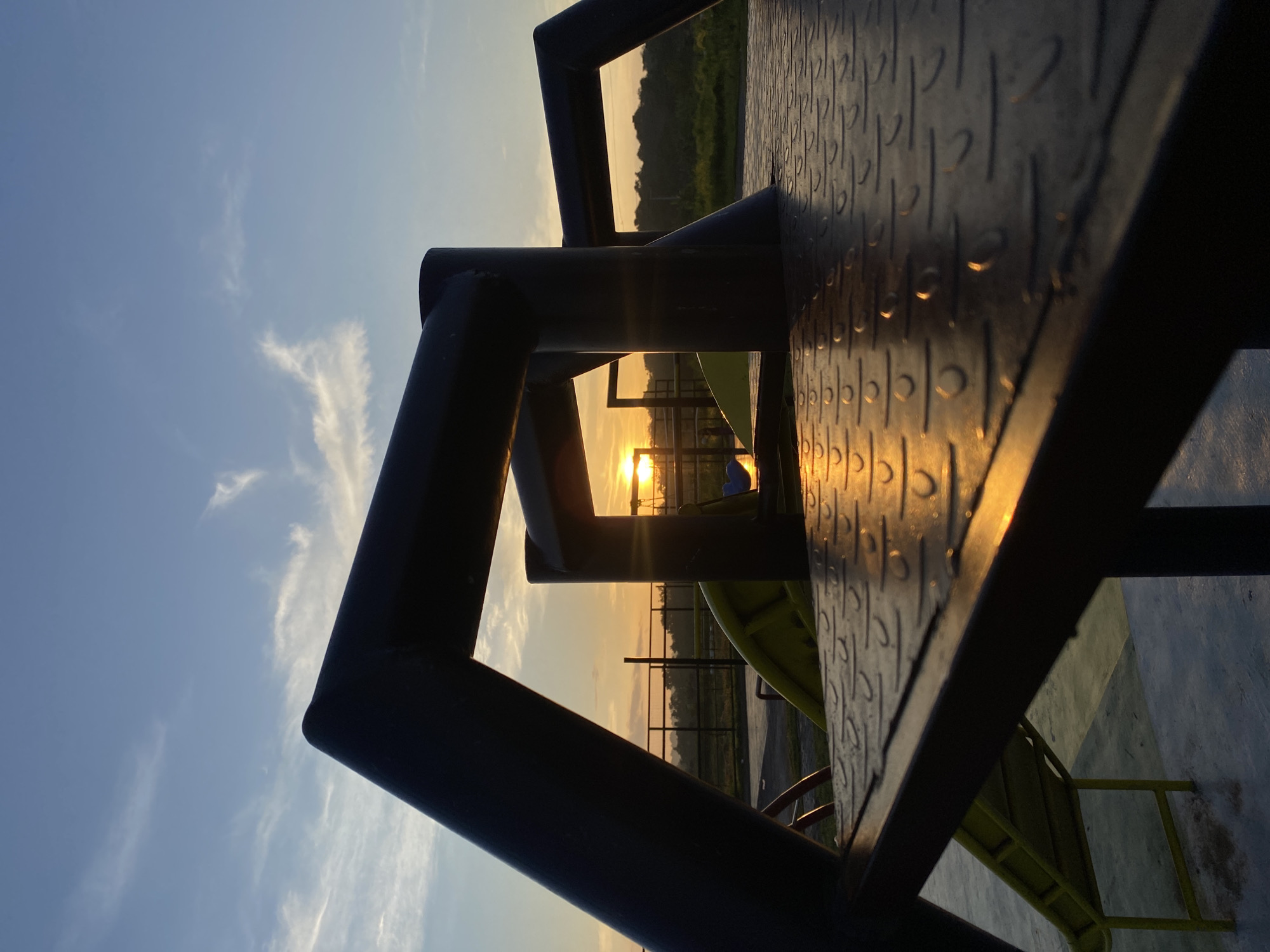 The image depicts a sunset at a playground in Davao City, Philippines. The foreground is dominated by the dark, metallic structure of playground equipment, possibly a climbing frame or part of a slide. The textured surface of a platform, catching the golden light of the setting sun, is prominently featured. In the mid-ground, a bright yellow slide is visible, partially obscured by the playground structure. Beyond the playground, a fence encloses the area, and a strip of green foliage marks the horizon. The sky is a blend of pale blue and soft orange hues, with wispy white clouds scattered across it, indicating the sun is low on the horizon. The overall impression is one of a serene evening at a recreational spot, likely a public park or a residential playground. The lighting suggests the time of day is either sunrise or sunset, with the warm glow of the sun creating long shadows and highlighting textures.