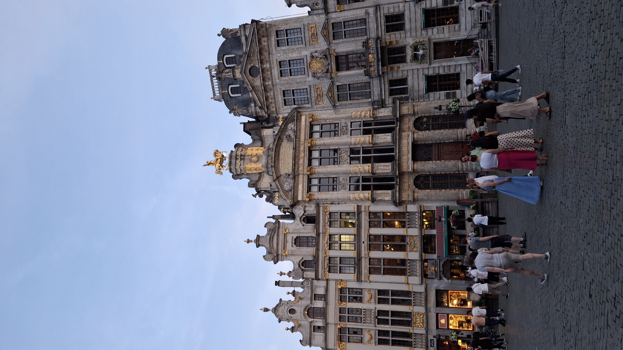 The image depicts a bustling city square with ornate, historical buildings lining the background. The architecture is characterized by intricate stonework, gilded accents, and statues adorning the facades. A prominent gold statue of a horse and rider stands atop one of the central buildings. Several people are visible in the foreground and midground, strolling across the cobblestone pavement. They are dressed in casual summer attire, suggesting a pleasant day. Some individuals are walking, while others are gathered in small groups, conversing. The sky is a pale blue, indicating it is either early morning or late afternoon. The overall scene conveys a lively, urban atmosphere, likely a tourist destination due to the grandeur of the architecture and the presence of many people.