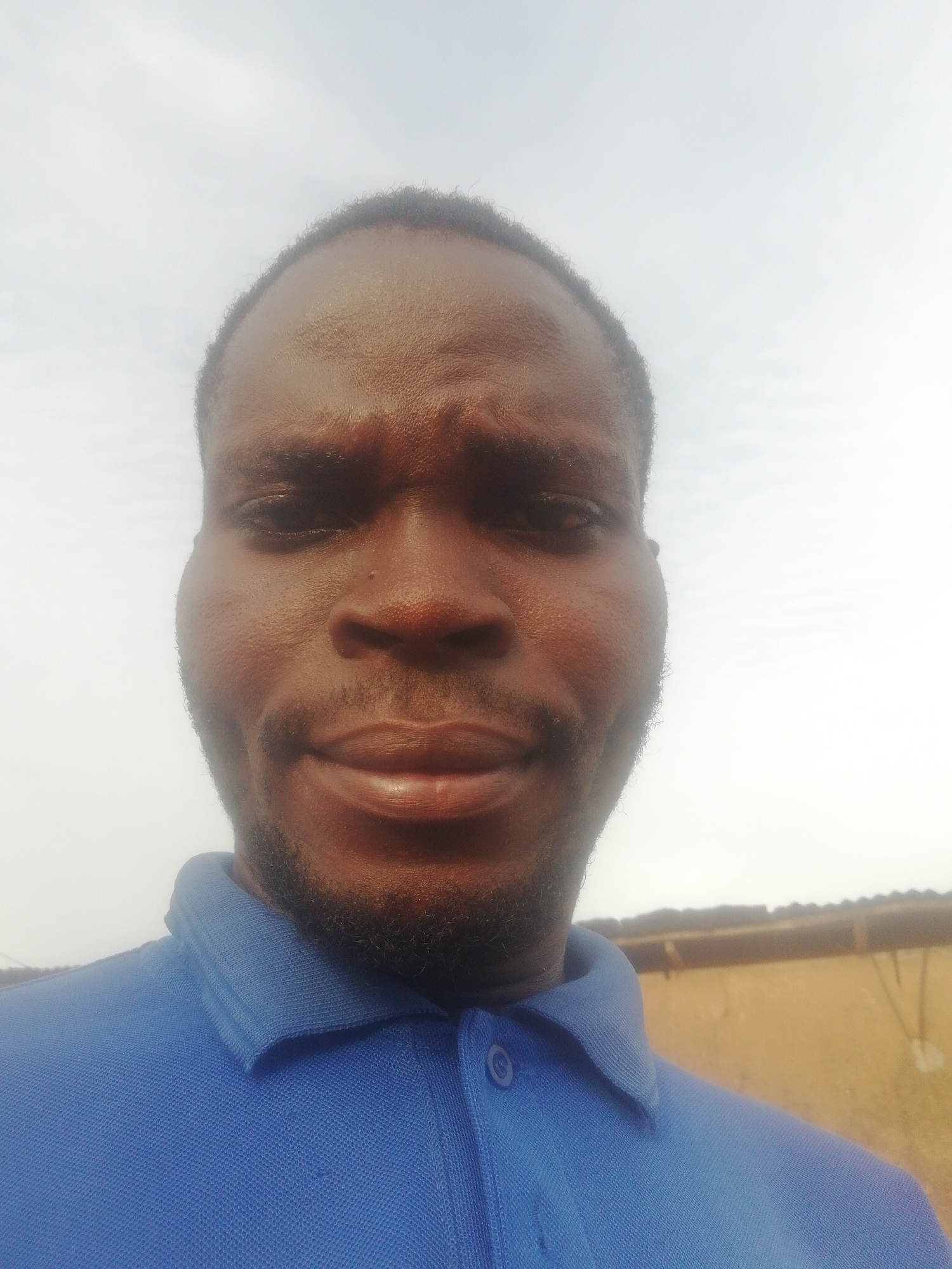 A close-up photograph shows a Black man wearing a blue collared shirt. He appears to be outdoors under a cloudy sky. A blurred background shows a building structure. The location is Unknown, Unknown.