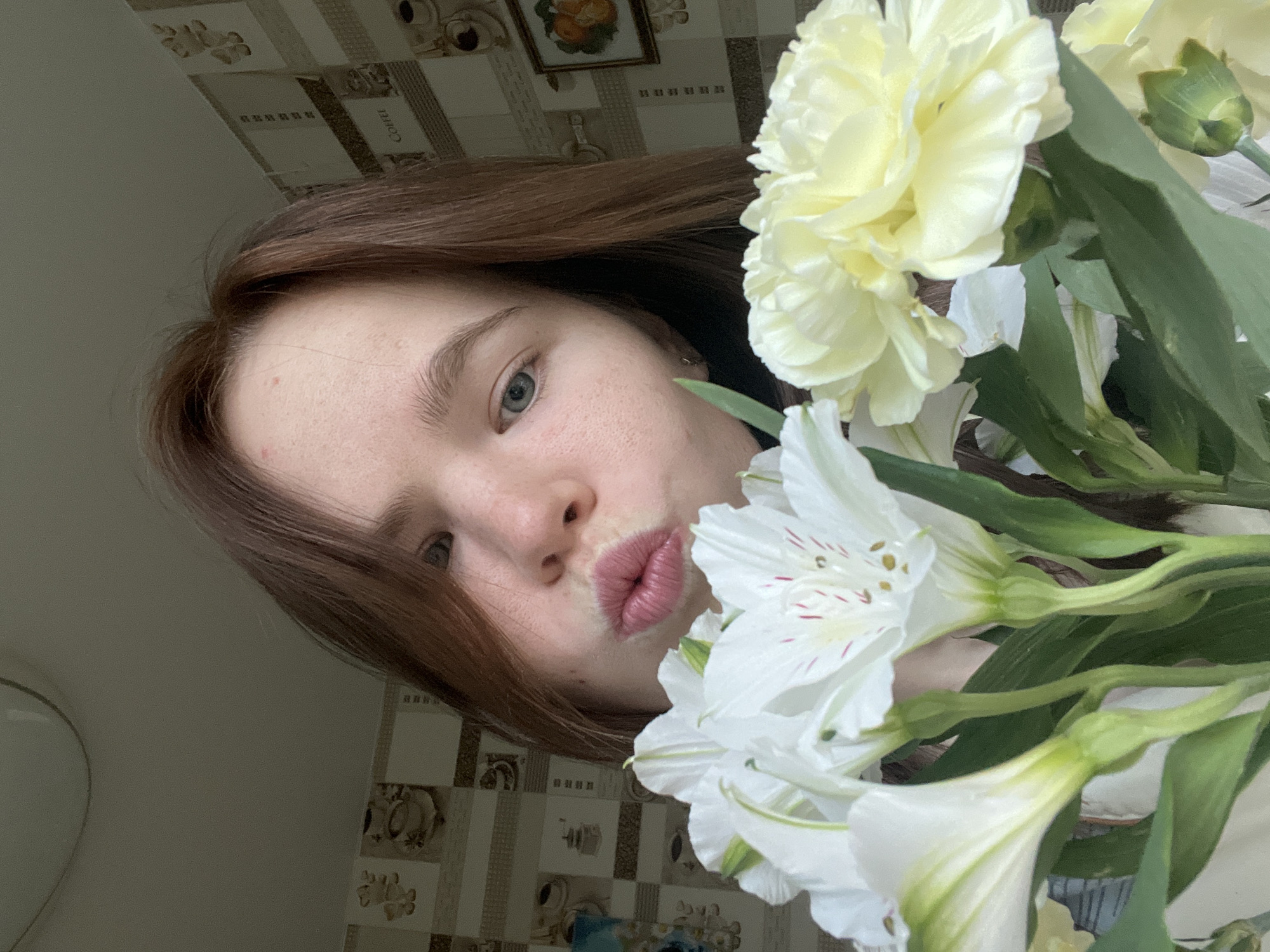 A young woman with light skin and reddish-brown hair poses for a selfie with a bouquet of flowers. She has pursed lips and looks directly at the camera. The bouquet contains white Alstroemeria and a large yellow flower, possibly a carnation. The background consists of a wall with decorative tiles featuring coffee-related imagery. The image doesn't offer sufficient context to ascertain if the photo was taken in Chaikovsky, Russia.