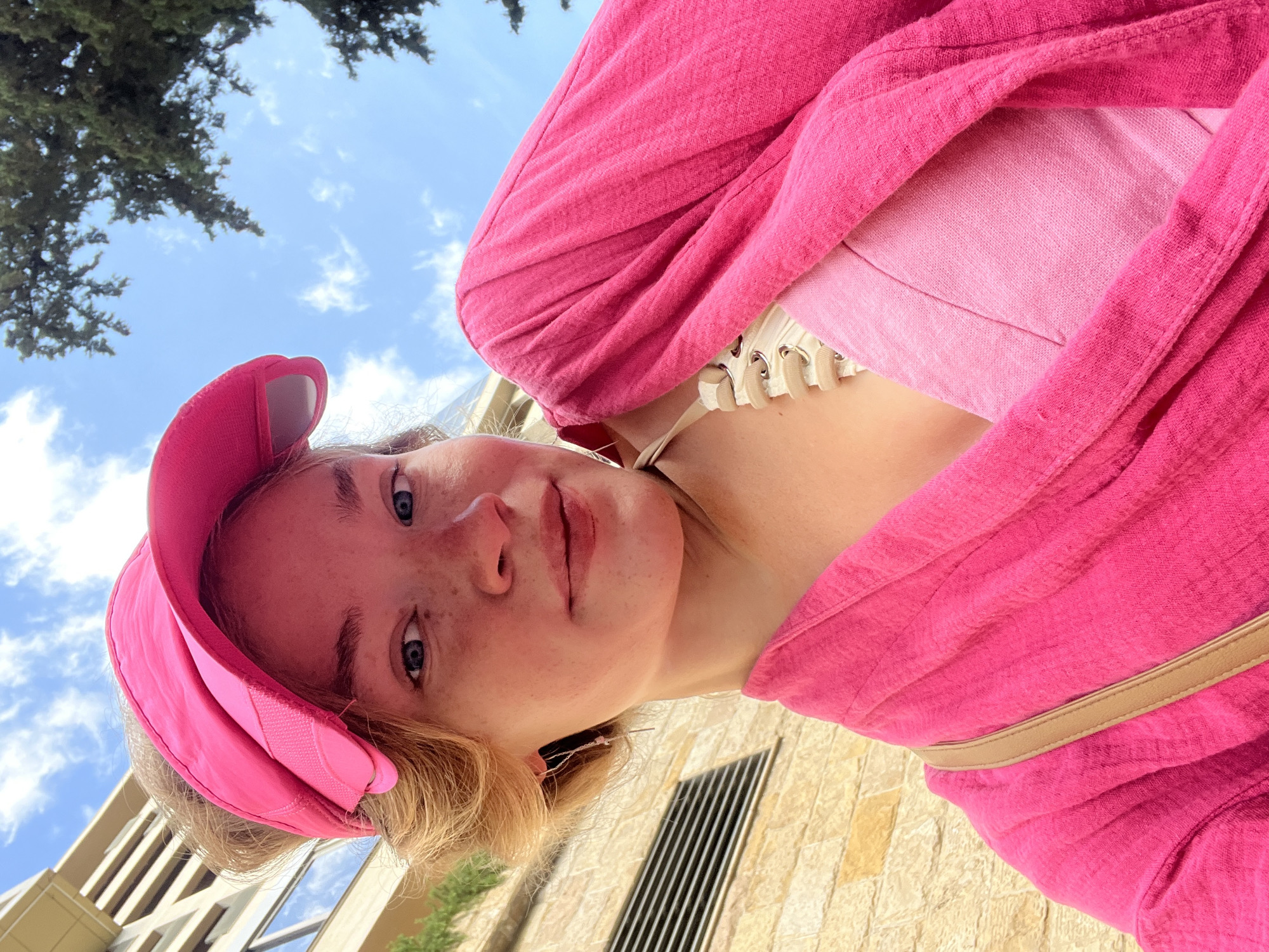 A young woman with fair skin, blue eyes, and freckles is captured in a low-angle, upward-facing photograph, taken outdoors. She is wearing a bright pink outfit, including a cap and a top that appears to be layered, with a lace-up corset detail visible at the neckline. The setting appears to be a public space in the city of Boreti, Montenegro, as evidenced by the presence of stone buildings and greenery. A section of clear blue sky with sparse white clouds is visible, as well as the top of a green tree. The woman is facing the camera with a neutral facial expression.