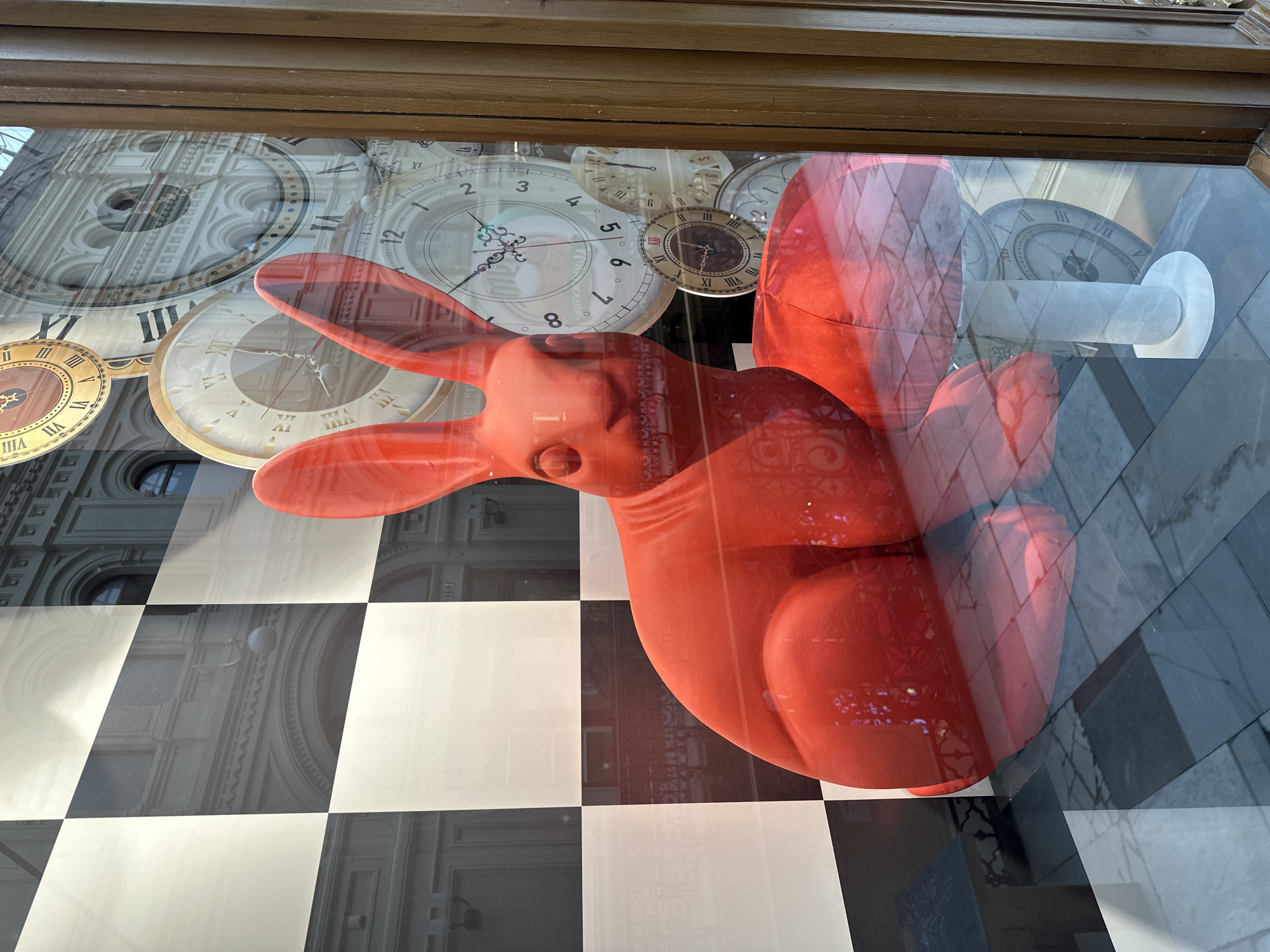 This is a close-up, eye-level shot of a red, stylized rabbit sculpture in a shop window display. The rabbit is large and appears to be made of a smooth, matte material. It is positioned centrally in the frame, seated on the floor, with its head tilted slightly to the left. Its ears are long and prominent, pointing upwards. To its right, a white, mushroom-shaped object on a pedestal is visible, with the rabbit's hindquarters partially obscuring it.
The background of the display is complex and layered due to reflections in the window. A black and white checkered pattern is prominent on the left side, suggesting a chess board motif. Behind and to the right of the rabbit, several large clock faces with Roman numerals are visible, some overlapping. These clocks are also reflections, showing architectural details of a building, likely across the street, with arched windows and ornate facades. The floor of the display appears to be tiled with a pattern of dark and light squares. A wooden frame of the shop window is visible on the far right. The overall scene is surreal and theatrical, evoking elements of "Alice in Wonderland."