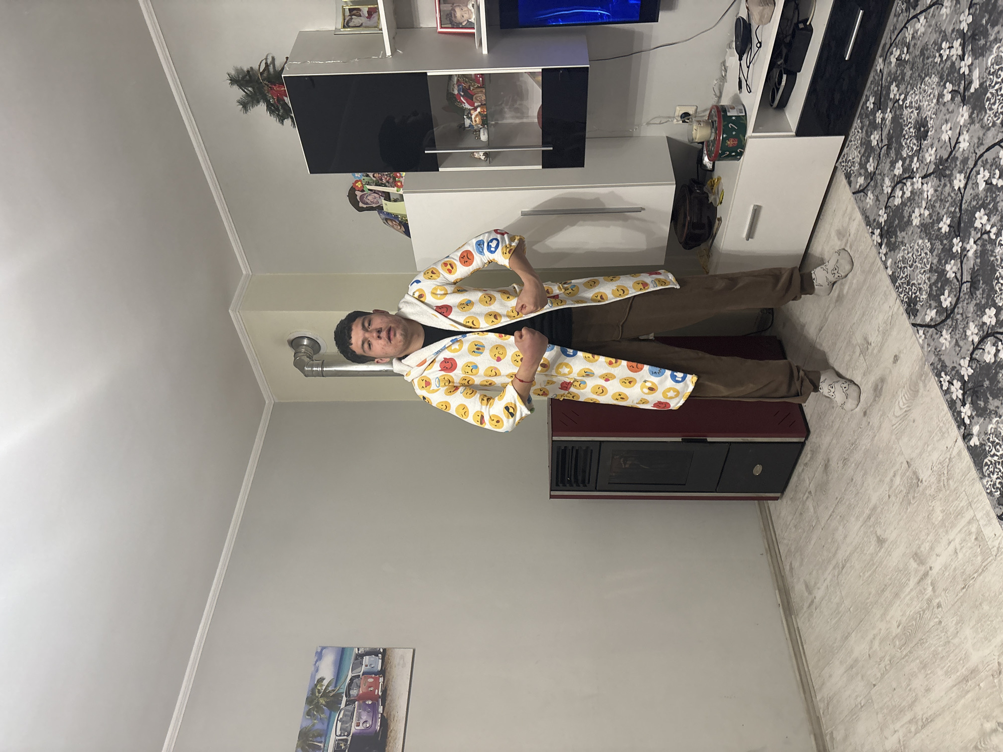 A man stands in a room. He wears a white robe patterned with emoji faces, brown pants, and white socks. Behind him are a dark red stove, a white entertainment center with black accents, a television, and a patterned rug. A painting hangs on the wall to his left. The location is a room in a residence. There is no indication it is in the city of Goritsa, Bulgaria.
