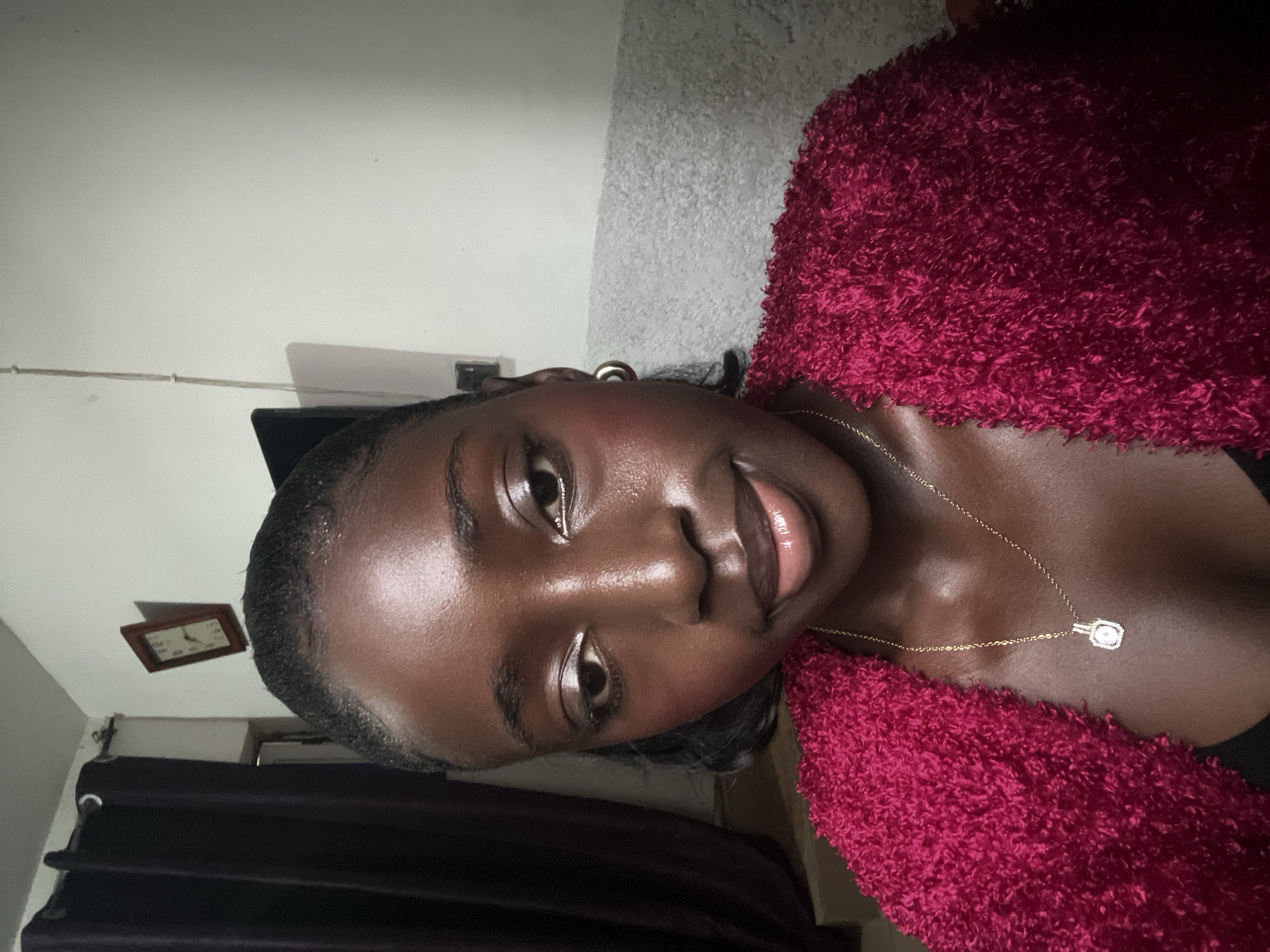 A close-up, slightly rotated portrait shows a dark-skinned woman with glossy skin and dark hair. She wears a gold necklace with a pendant and a textured burgundy top. The background consists of a white wall and dark curtains. A small framed object hangs on the wall in the background. The location is likely an interior space, but details specific to Abuja, Nigeria, are not discernible.