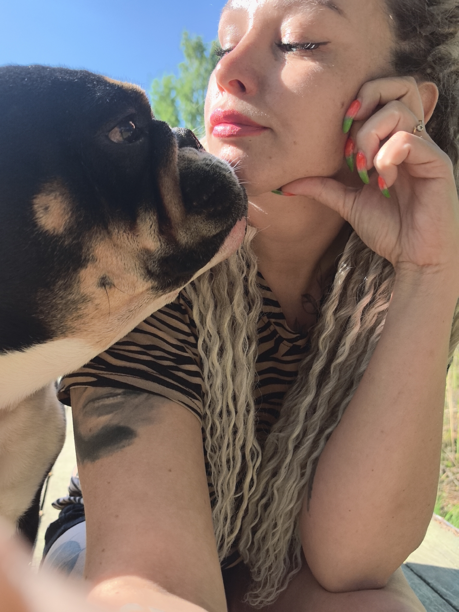 A woman with light skin, long, curly blonde hair, and red lipstick is interacting with a black and tan dog. The woman's eyes are closed, and she has her hand to her chin. She wears a shirt with a black and brown tiger print and has tattoos on her arm. Her fingernails are decorated with a green and red design. The dog's face is close to hers, and its eye is visible. The background includes green trees and a blue sky. The exact location within the city of Noviy, Russia, cannot be determined from this image.