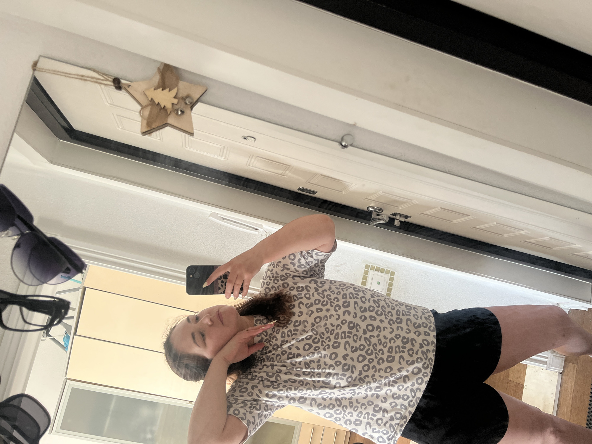 In a full-length mirror, a woman with brown hair tied back takes a selfie. She is wearing a gray t-shirt with a black leopard print and black shorts. Her left arm is bent and resting on her head, while her right hand holds the phone and her left hand is touching her face. In the reflection of the mirror, a white door with wood paneling is visible, along with a wooden star ornament hanging from a string on the door. To the left of the door, in the background, are beige cabinets. The woman's pose and the selfie suggest a casual moment, possibly at home, as suggested by the domestic setting reflected in the mirror. The image does not contain any information or visual cues that would allow for its location to be identified as Chelyabinsk, Russia.
