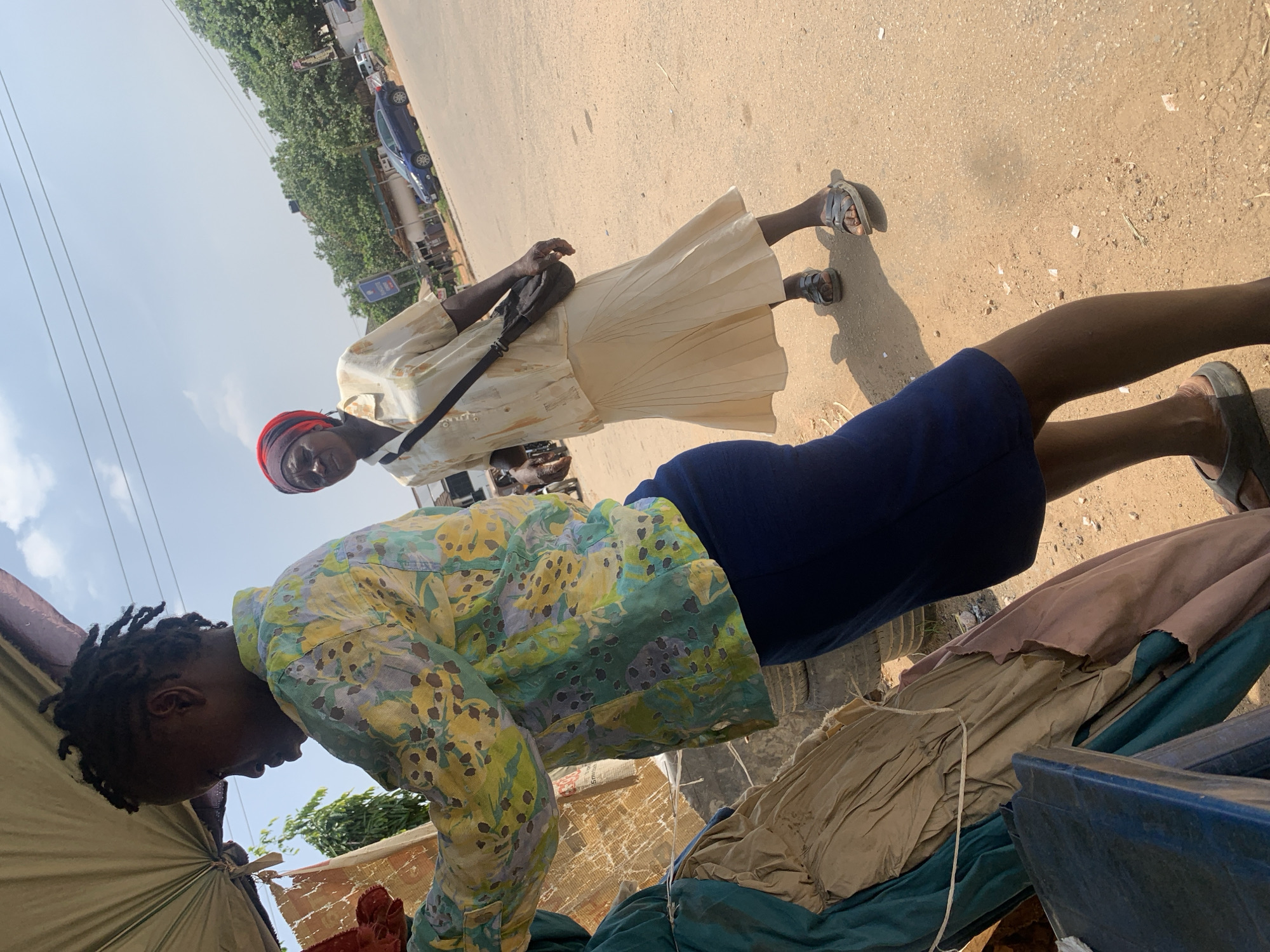 The image captures a street scene, likely in Abuja, Nigeria, under a bright, partly cloudy sky. In the foreground, a person viewed from the side is wearing a colorful, patterned long-sleeved shirt and a dark blue skirt. They are standing next to a display of folded fabrics, which appear to be rough textiles or tarpaulins in shades of green, beige, and pink. The fabrics are tied with rope and appear weathered and perhaps for sale or storage.
Behind this person, another individual is walking away from the camera, wearing a light-colored, short-sleeved button-up shirt, a cream-colored pleated skirt, and a red headwrap. A dark bag is slung across their right shoulder. This person is walking on a dusty, unpaved road.
The background reveals more of the environment: a row of trees lines the opposite side of the road, and beyond them, buildings and structures can be seen. Several vehicles, including a blue car, are present on the road, suggesting it's a public thoroughfare. The overall impression is of an active street market or commercial area in an urban setting. The lighting indicates it is daytime, with shadows cast by the people and objects.