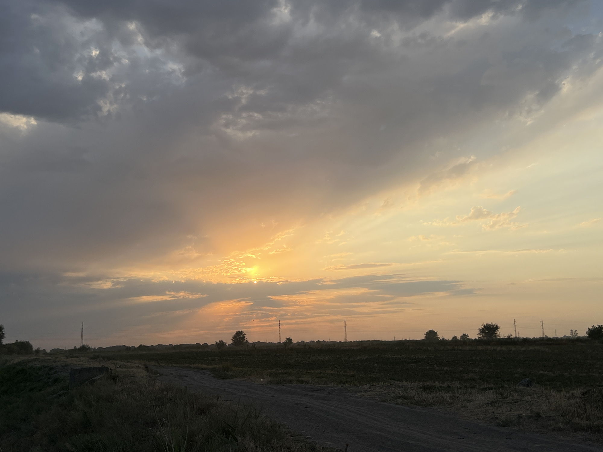 A wide shot captures a rural landscape during a sunset. The sky dominates the upper half of the image, with a blend of dark, heavy clouds on the left and lighter, more scattered clouds illuminated by the setting sun on the right. The sun itself is a bright, concentrated orb peeking through the clouds, casting a warm orange and yellow glow. Rays of sunlight extend outwards, creating streaks of light across the sky.
The horizon is marked by a flat, dark field that appears to be agricultural land, with some patches of dry grass. A dirt road or path cuts through the foreground, leading towards the horizon. Several scattered trees and utility poles are visible in the distance, silhouetted against the bright sky. The overall mood is serene and atmospheric, characteristic of a sunset in a rural setting.