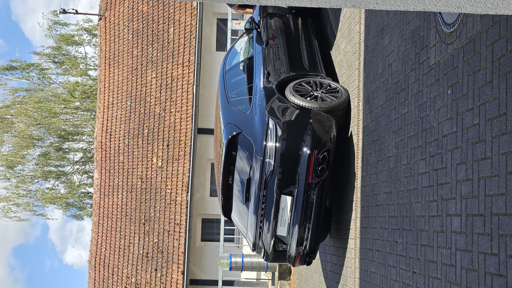 An eye-level, slightly low-angle, medium shot shows the rear of a black Porsche Panamera wagon parked on a paved surface in front of a building with a red tiled roof. The car is the primary subject, positioned centrally and facing towards the right of the frame. Its sleek, dark exterior is highly reflective, mirroring the sky and surrounding environment. The vehicle features prominent quad exhaust pipes at the rear, a spoiler, and a license plate bracket with "PORSCHE APPROVED" signage. To the left of the car, a metal railing encloses some cylindrical objects, possibly barrels, and a white building with windows is visible. The ground is paved with dark grey bricks laid in a herringbone pattern, with a metallic grate running horizontally across the scene in the middle ground. Above the building, a bright sky with scattered white clouds is visible, partially obscured by the green foliage of a tree. The overall lighting suggests a sunny day. No people are clearly visible in the frame.