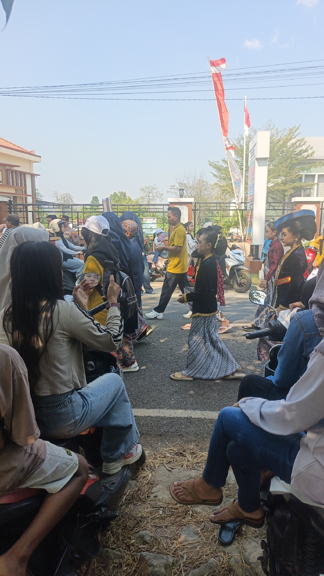 This is a full-shot, vertical, outdoor photograph taken on a sunny day. The scene appears to be a street, possibly during a parade or procession, given the presence of people in traditional clothing and a crowd gathered. In the foreground, several people are seated on what appear to be motorcycles, facing away from the camera. Their legs and lower torsos are visible, with one person wearing blue jeans and a light-colored jacket, and another person wearing light brown shorts and a light brown shirt. The ground is covered with dry grass and some scattered debris.
In the mid-ground, a group of people are walking along the street. A young person in traditional attire, consisting of a dark jacket and a patterned skirt, is walking towards the right of the frame. Behind them, other individuals are seen, some in casual clothing and others in traditional garments. A man in a yellow t-shirt and dark pants is walking in the opposite direction. Motorcycles are parked along the side of the road, and a few people are riding them.
In the background, a building with red-tiled roofs is visible on the left. A tall white pole with a sign is on the right, and two Indonesian flags are flying. The sky is clear and blue, with a few power lines crisscrossing overhead. The overall atmosphere suggests a public gathering or event.