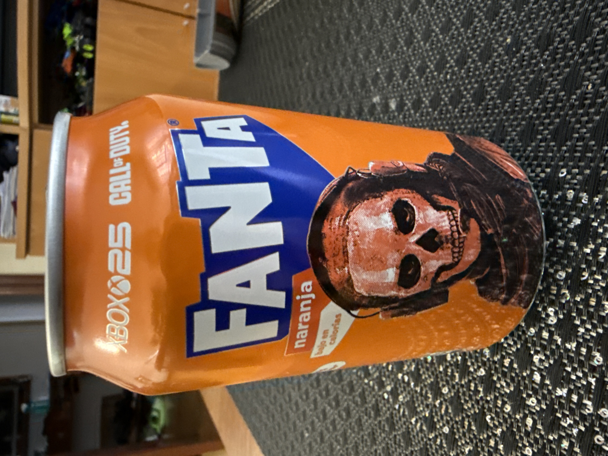 A close-up, eye-level shot shows an orange Fanta soda can with a Call of Duty and Xbox 25th anniversary collaboration design. The can features the Fanta logo in large blue letters, the Xbox and Call of Duty logos at the top, and a graphic of a skull wearing headphones. A small orange label reads "naranja" and a white label below it reads "bajo en calorias." The can is placed on a dark gray textured surface, possibly a table, with a blurred background showing wooden furniture and shelves containing various items, including what appears to be toys. The lighting suggests an indoor environment. There is no indication of this image being taken in Pamplona, Spain.