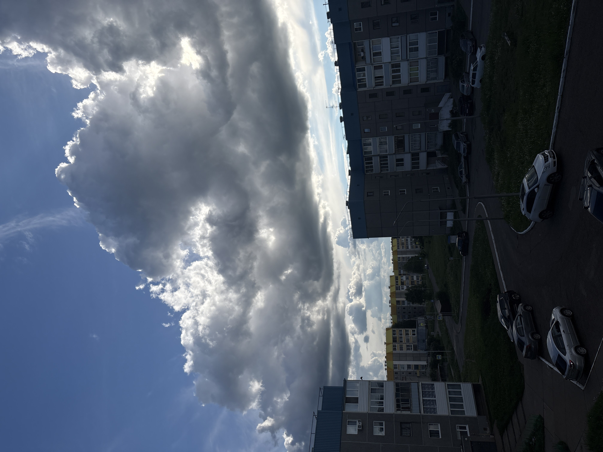A low-angle view shows a residential area with apartment buildings under a partly cloudy sky. The photo is slightly rotated, with the sky to the left and buildings to the right. The clouds are large and billowy, ranging from light gray to darker storm clouds, with sunlight breaking through in some areas. Several multi-story apartment buildings are visible, arranged in rows and varying in color. A street runs between the buildings, with parked cars lining the roadside. The scene is from Markovskiy, Russia, evidenced by the architecture and style of the buildings. There are no people visible.