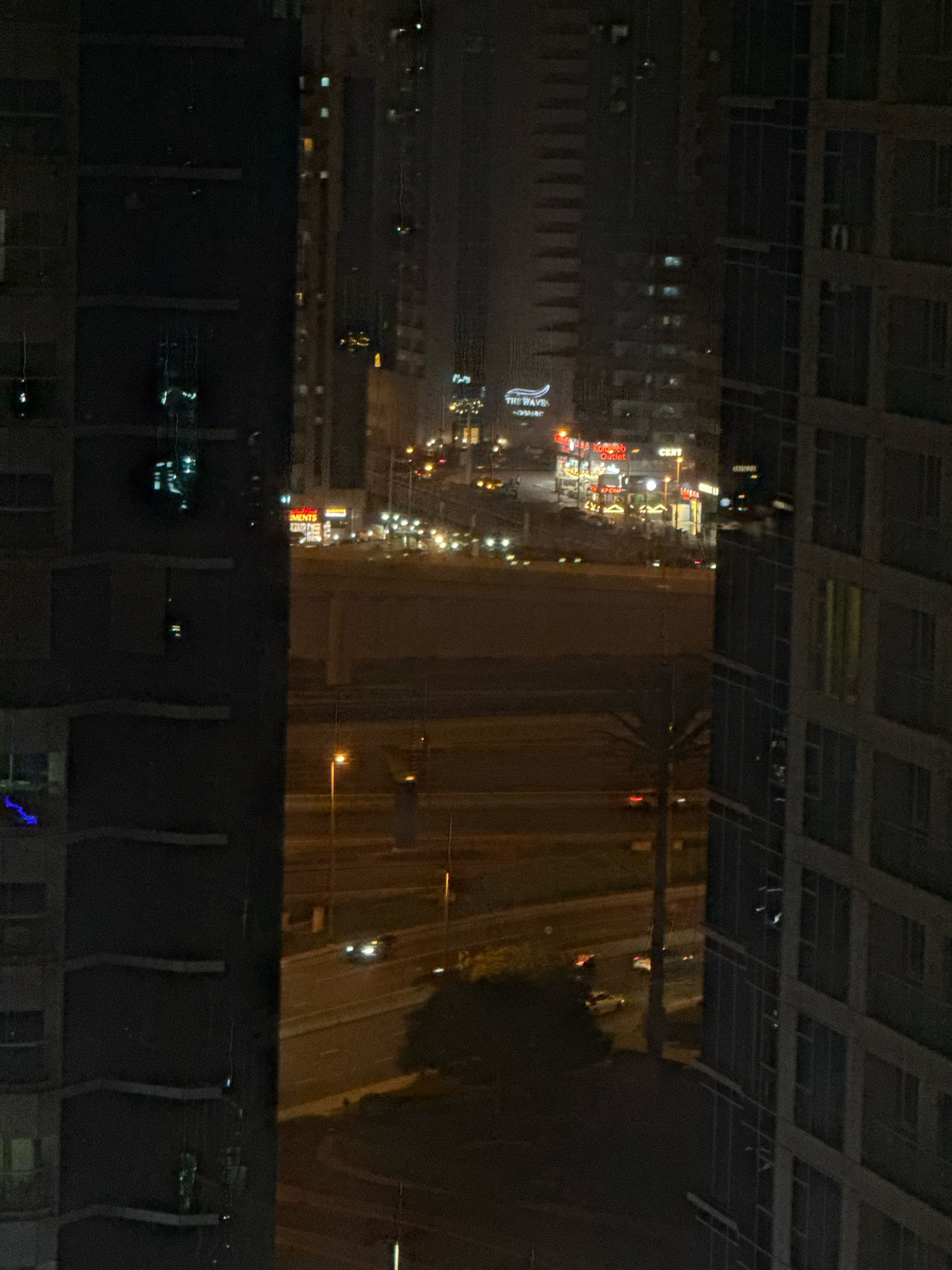 The image depicts a nighttime view of a cityscape in Dubai, United Arab Emirates, likely from a high vantage point. The foreground is dominated by the dark, imposing facades of two modern high-rise buildings, one on the left and another on the right, with their windows reflecting faint light. Between these buildings, the urban landscape unfolds. In the mid-ground, a busy street or highway is visible, illuminated by streetlights and the headlights of passing vehicles. Several cars are visible on the road, moving in both directions. Beyond the road, a cluster of commercial buildings and shops are lit up, displaying signs with Arabic text and logos, including one that reads "THE WAVE" with a distinctive wave graphic. Further in the background, more high-rise buildings stretch into the dark, hazy sky, their windows appearing as scattered points of light. The overall impression is one of a vibrant, active urban environment at night, characterized by modern architecture and constant movement. There are no visible people in the frame.
