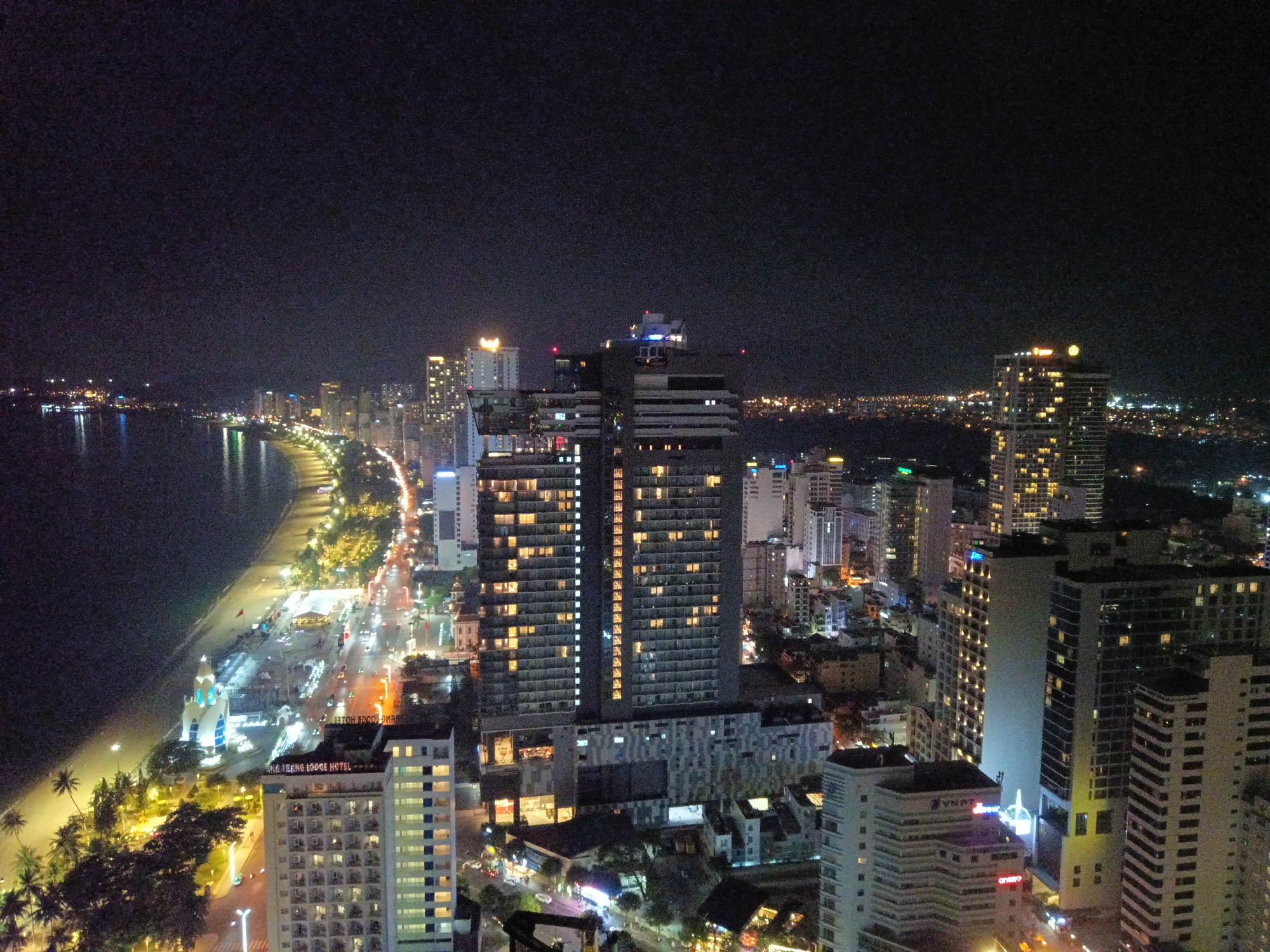 This is an aerial view of Nha Trang Ward, Vietnam at night. A coastline with sandy beach is lit by streetlights and building lights. Buildings of varying heights dominate the city skyline, illuminated with numerous lights. Some buildings are particularly tall and distinctively designed. The night sky is dark and mostly featureless. The overall scene is characterized by urban development along the coast.