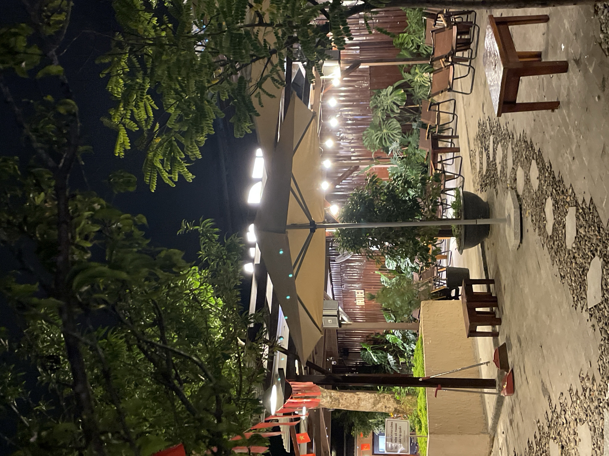 The image depicts an outdoor seating area at night, likely a cafe or restaurant, located in Lâm Viên - Đà Lạt, Vietnam. The scene is illuminated by string lights hanging above a large, beige umbrella. Several wooden tables and chairs are arranged on a paved patio, which is interspersed with gravel and decorative stones. Lush green plants, including large-leafed varieties, are visible in pots and the background. In the foreground, a broom and dustpan are leaning against a planter. A small wooden bench and a low wooden table are also present. The background features a rustic wooden fence and some signage in Vietnamese. The overall atmosphere suggests a tranquil evening setting.
