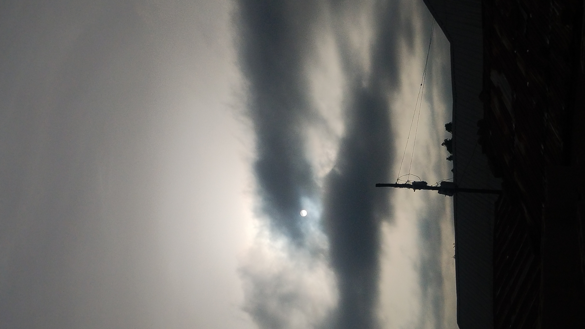 The image shows an overcast sky in Jos, Nigeria. The sun is visible as a bright white orb, obscured by layers of gray and dark clouds. In the foreground, silhouetted against the sky, are utility poles with wires and a dark building structure on the right. The overall scene conveys a sense of gloominess.