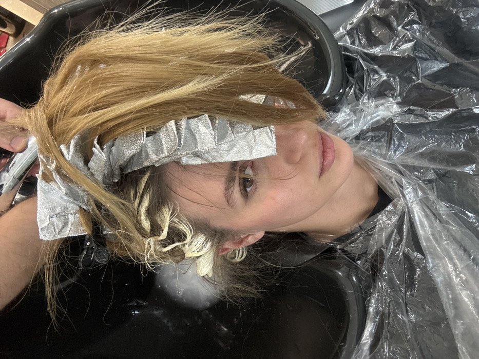 A young adult female is depicted from a high angle, looking directly forward, while undergoing a hair treatment in a hair salon. Her head is positioned within a black hair washing basin, which contains some white, foamy residue. Sections of her blonde hair are wrapped in multiple silver foils, indicating a highlighting or coloring process. Other strands of hair, particularly on the left side near her scalp, show evidence of a light-colored product application. A portion of her loose blonde hair hangs over her right eye. She is wearing a clear plastic protective cape over her torso. An unseen individual's hand is visible at the top center, manipulating a section of her hair and holding a chrome-plated metal object, likely part of a salon faucet or spray hose. The specific location within the city of Tbilisi, Georgia, cannot be determined from the image.