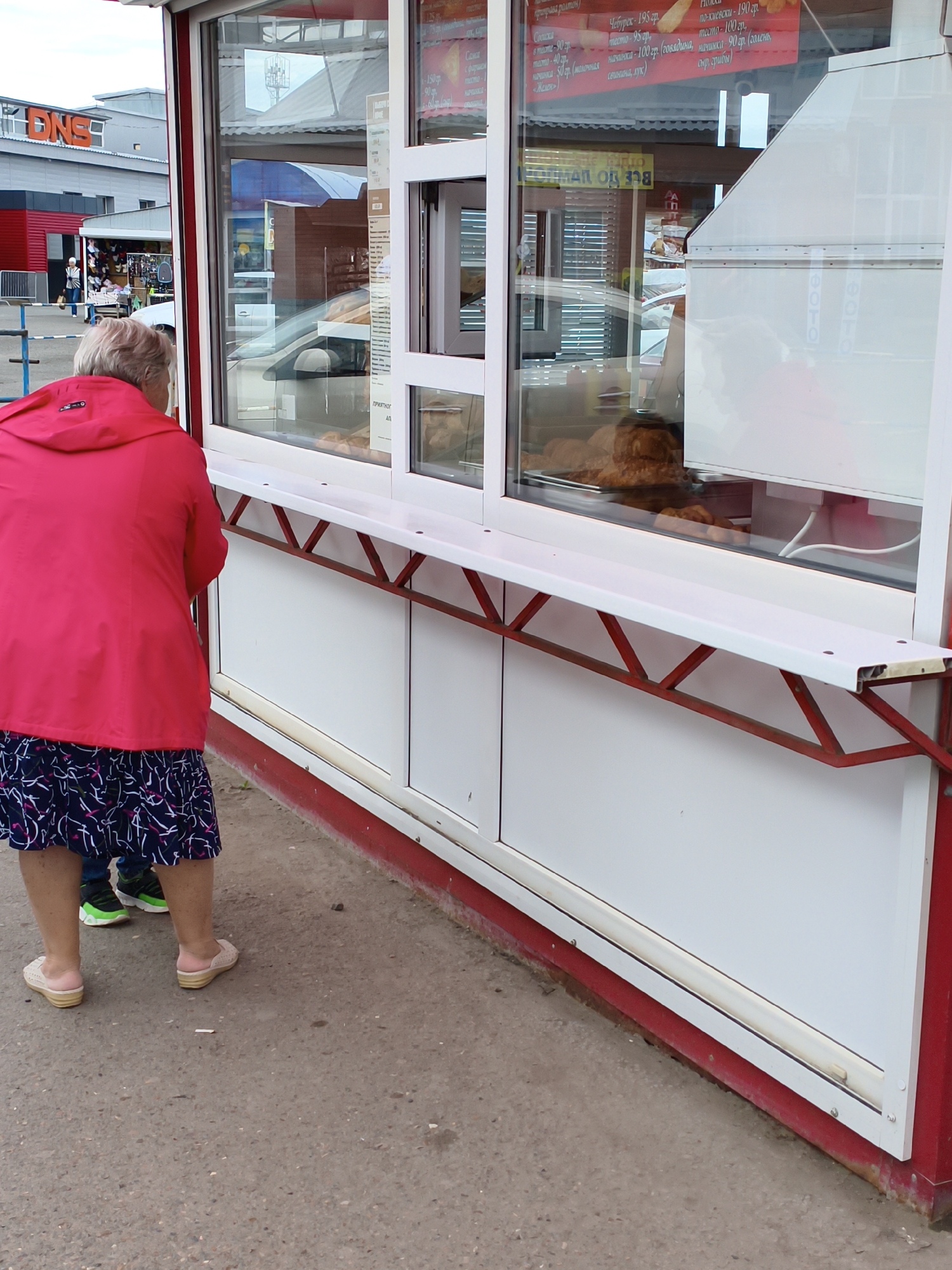 An elderly woman wearing a bright pink jacket and a floral skirt stands with her back to the camera, facing a small food kiosk. The kiosk has a large glass window displaying baked goods. Reflected in the window are buildings and the sky. A younger person, wearing athletic shoes, stands slightly behind and to the side of the woman. The scene is outdoors, with a paved ground surface and a red and white structure of the kiosk. The overall impression is a casual, everyday scene in a public space. Based on the signage visible in the background, the location is likely a commercial area.