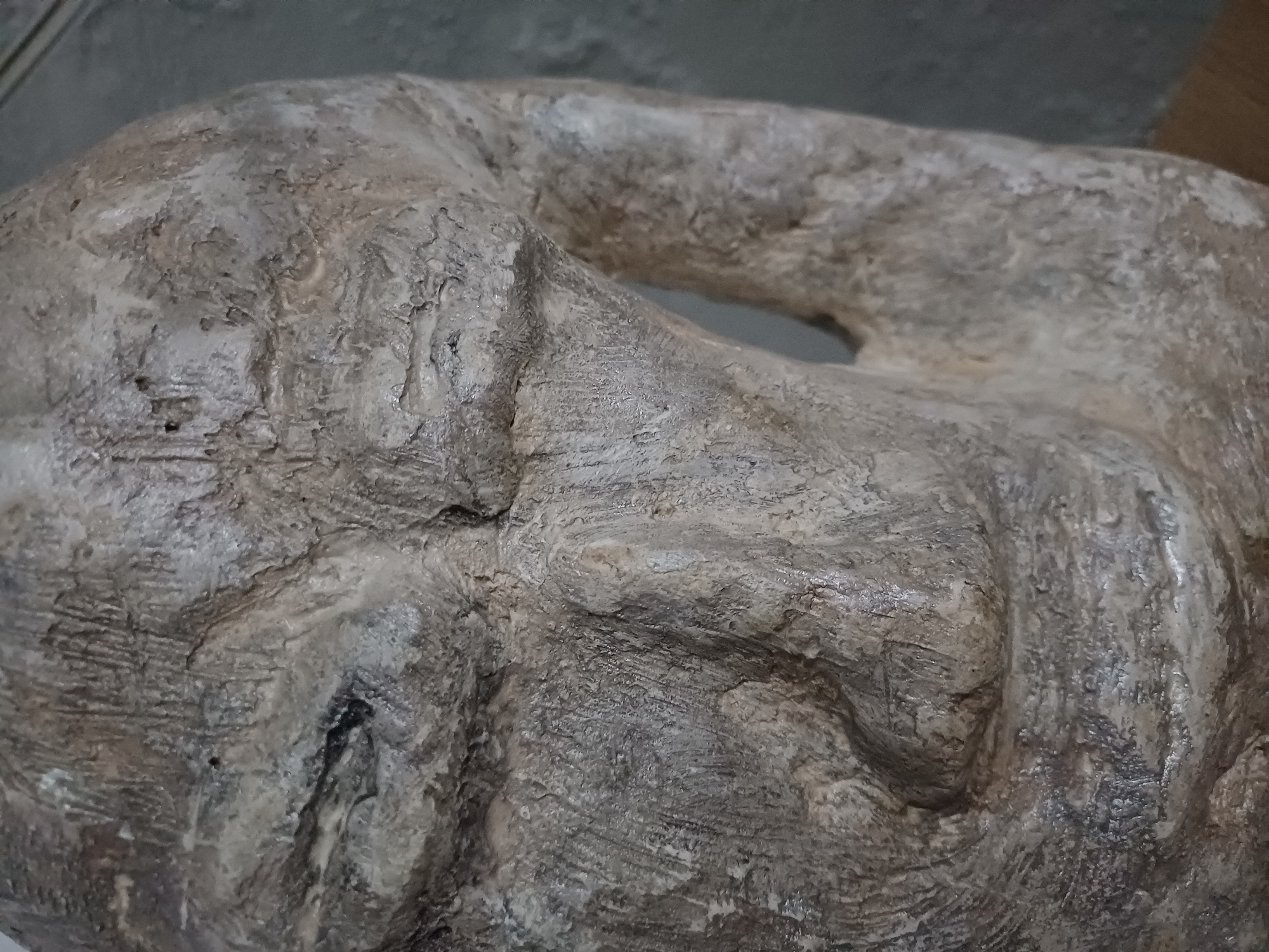 The image shows a close-up of a weathered, textured sculpture, possibly made of clay or stone, in the city of Sulymivka, Ukraine. The sculpture appears to be a stylized representation of a face, with prominent features such as an eye socket and nose carved into the rough surface. The material is a light grayish-brown, with variations in tone and texture that suggest age and exposure to the elements. The lighting is diffused, casting subtle shadows that accentuate the sculpted forms. The background is out of focus, showing a neutral gray wall.