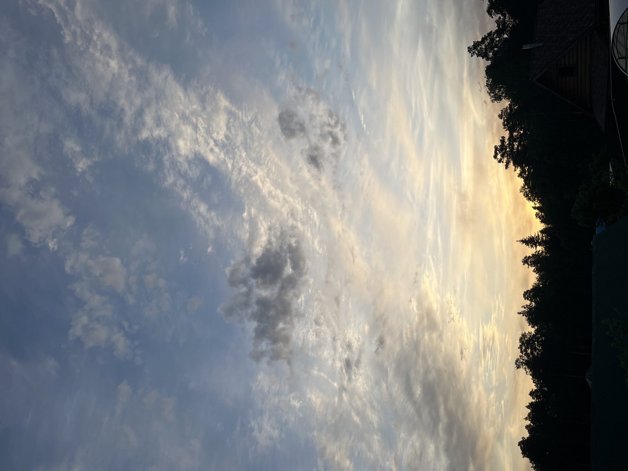 The image shows a view of the sky at sunset, likely in or near Chaikovsky, Russia, given the user's location. Wispy clouds are scattered across the blue sky, with a patch of darker gray clouds. On the right, a line of trees and the roof of a building are silhouetted against the yellowish light of the setting sun.