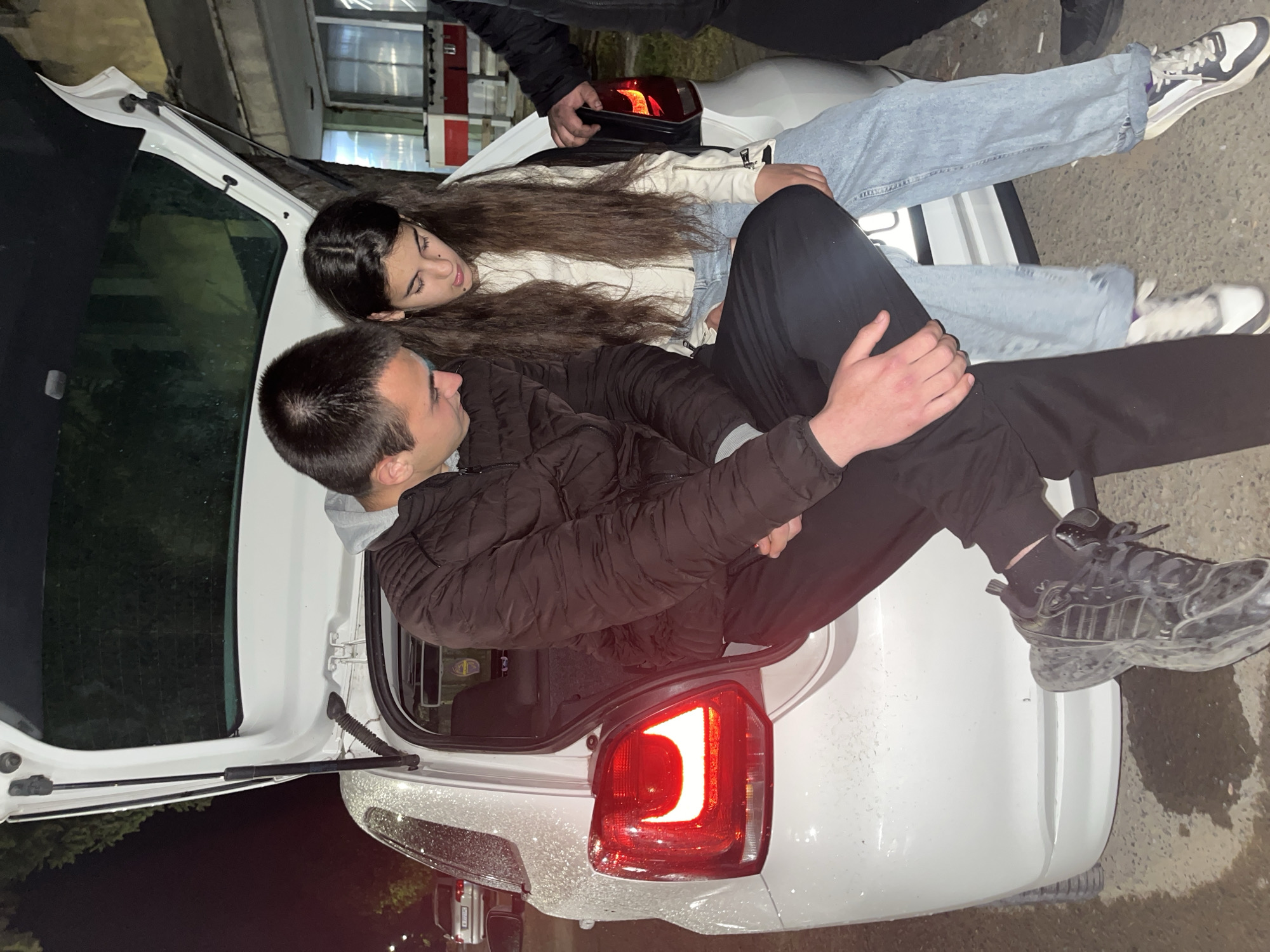 The image shows a group of young people gathered around the open trunk of a white car. A young woman with long brown hair and wearing jeans and a light-colored jacket is reclining in the trunk, interacting with a young man wearing a black puffy jacket and black pants, who is also positioned in the trunk. The car's taillight is illuminated. Another person is partially visible with their legs and sneakers on the left edge. The setting appears to be outdoors, at night, with an urban backdrop of buildings. The car is parked on what seems to be a wet surface. The location is identified as Lozarevo, Bulgaria.