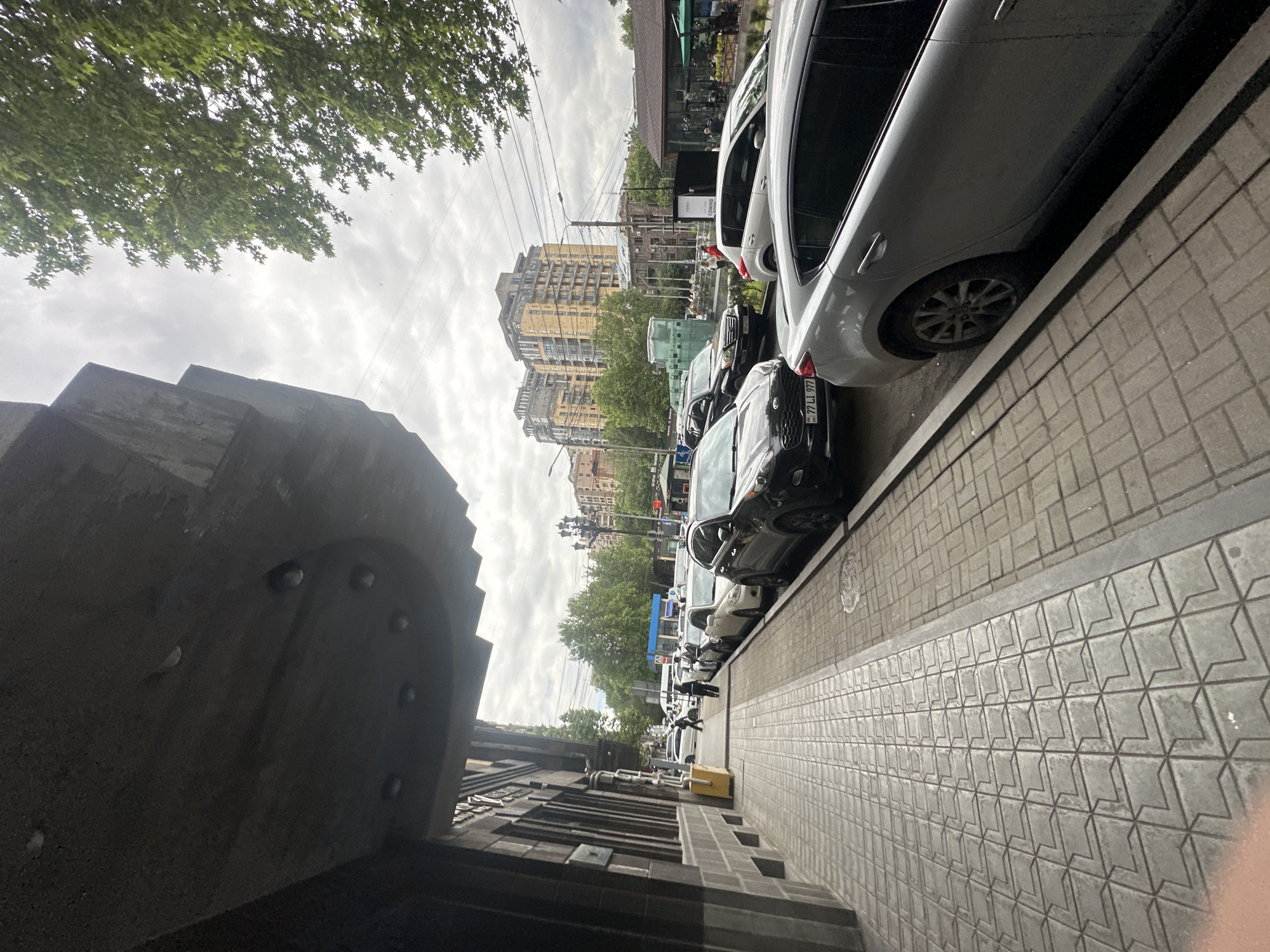 The image depicts a street scene in Yerevan, Armenia. The camera is positioned at ground level, looking along a sidewalk towards a street lined with parked cars. Buildings are visible in the background, and trees line the street. The sky is overcast. The architecture features elements of Soviet-era construction. The sidewalk is paved with patterned tiles. The cars parked on the street include a white hatchback near the foreground and other vehicles of various colors and models further down the street. Power lines are visible in the sky. A section of a building with distinct architectural features and a rounded element is visible on the left side of the frame.