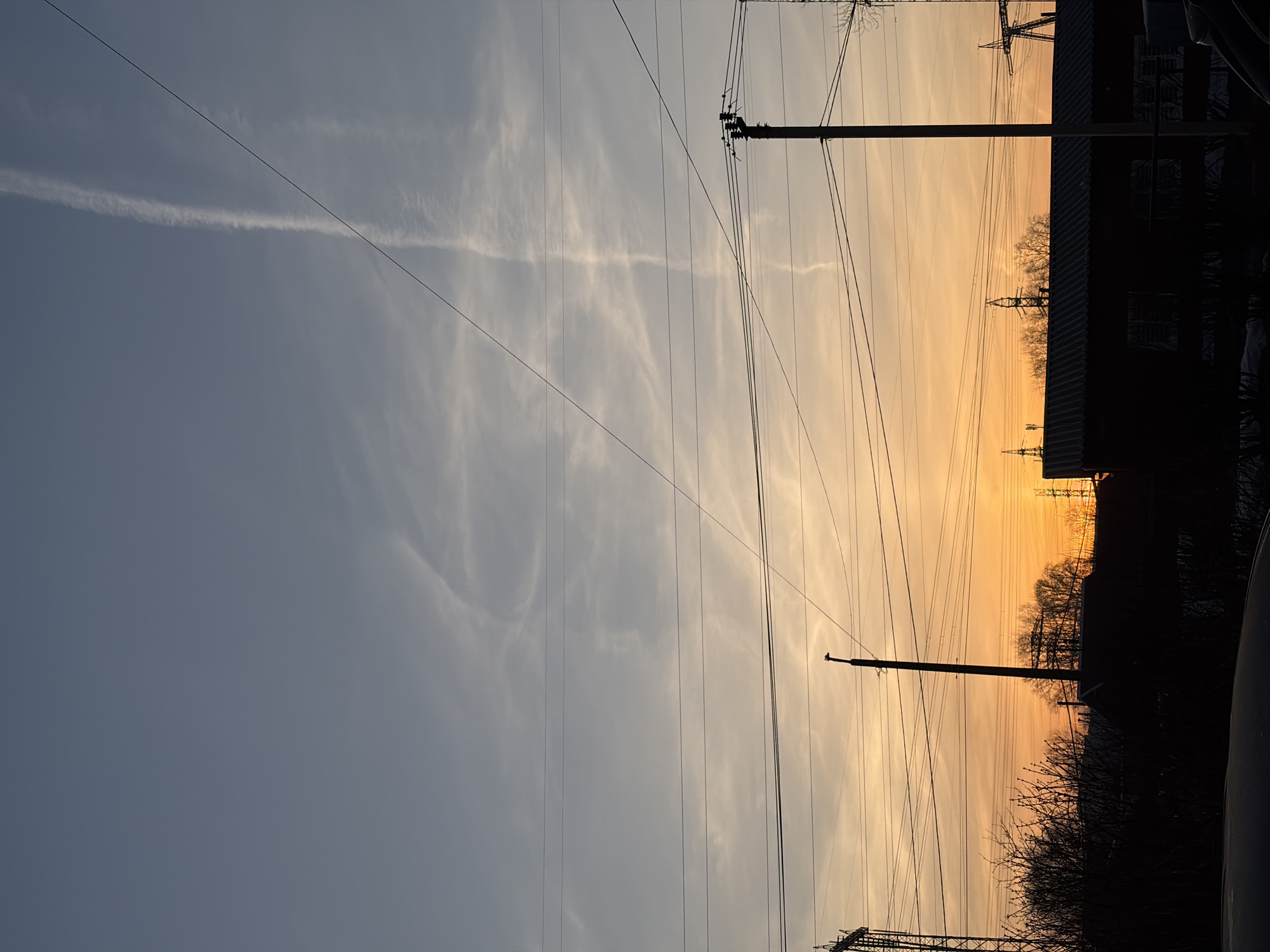The image depicts a sunset sky in the city of Chaikovsky, Russia. A network of power lines and utility poles runs across the frame. The sky transitions from a pale blue-gray to a warm orange-yellow near the horizon. Cirrus clouds are present in the sky, with a distinctive contrail visible. Silhouetted against the sunset are buildings, trees, and other infrastructure. The image appears to be taken at ground level.