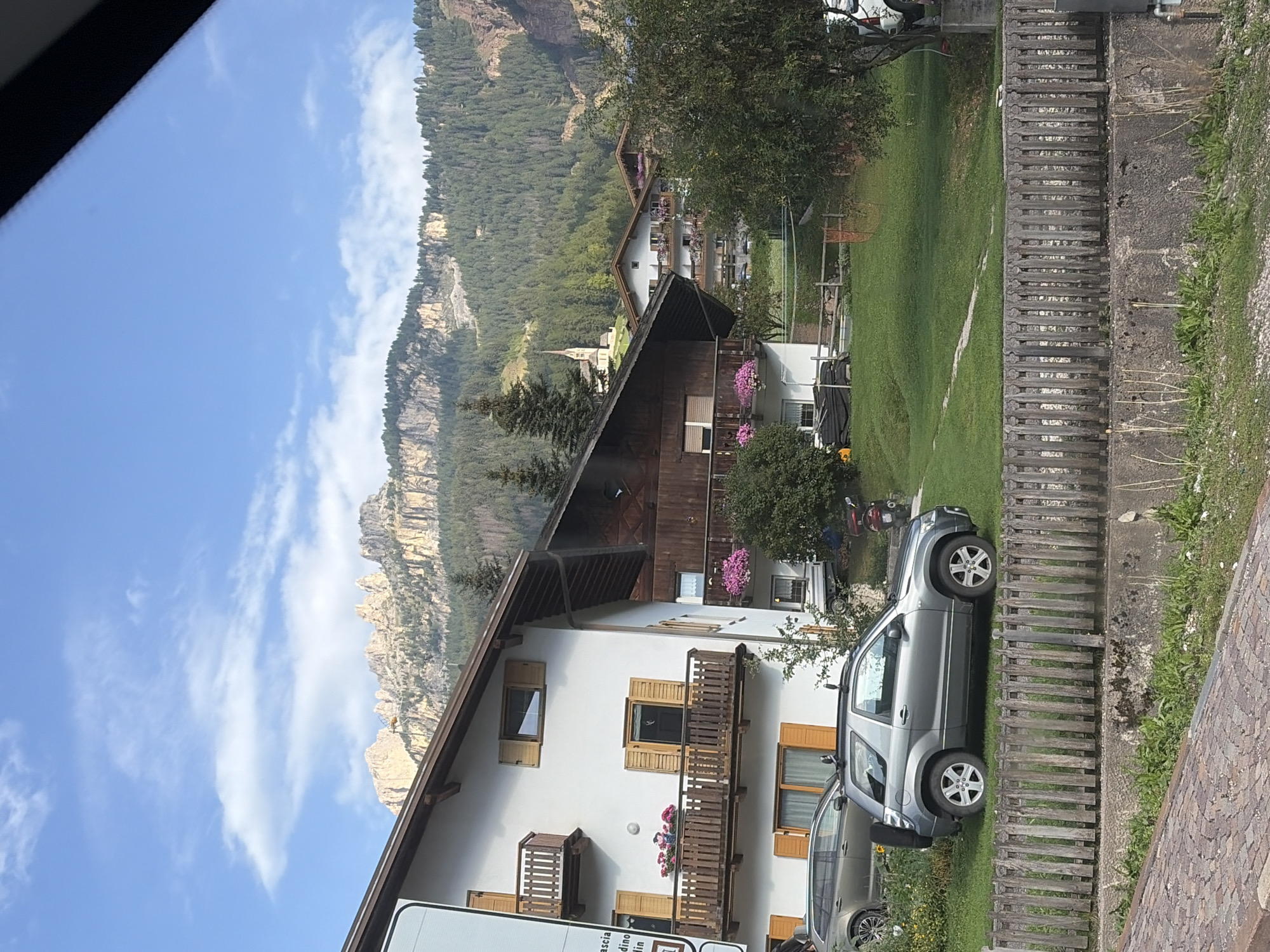 The image depicts a view of a village nestled in a mountainous region, likely in Vigo di Fassa - Vich, Italy, given the architectural style and mountainous backdrop. In the foreground, a wooden fence runs horizontally, separating a cobblestone path on the left from a grassy area on the right. Two cars are parked on the grassy area, one a silver SUV and another a darker grey SUV. Beyond the cars, several houses are visible. The house on the left is white with dark wooden balconies and shutters. The house on the right is primarily dark wood with balconies. A small church with a steeple is visible further up the hill, surrounded by trees. The background is dominated by steep, rocky mountains covered in dense green coniferous forest. The sky is blue with scattered white clouds. The overall impression is a serene, picturesque mountain village scene.