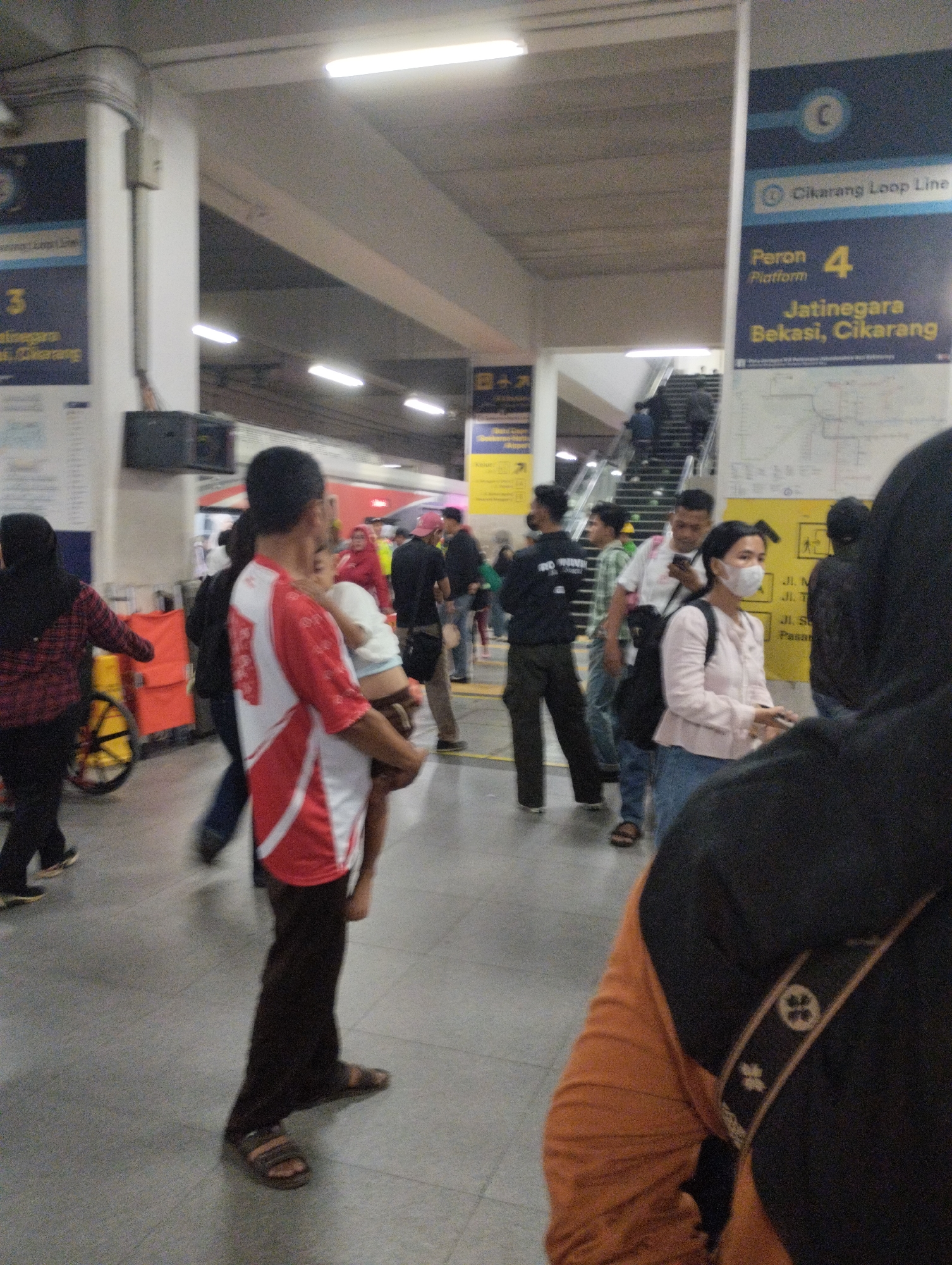 The image depicts a bustling indoor environment, likely a train station, with a crowd of people. In the foreground, a man in a red and white shirt is holding a child. He is wearing dark pants and sandals. To his left, a woman in a black hijab and red and black checkered shirt is visible. Behind them, other individuals are seen walking and standing, some with bags. Signs are visible in the background, including one that reads "Cikarang Loop Line" and "Peron 4" in English, indicating a platform number. The flooring is tiled, and fluorescent lights illuminate the space from above. The overall atmosphere is that of a busy transit hub with people on the move. The specific location in South Jakarta, Indonesia, is not directly ascertainable from the visual information alone, but the Indonesian signage suggests the country.