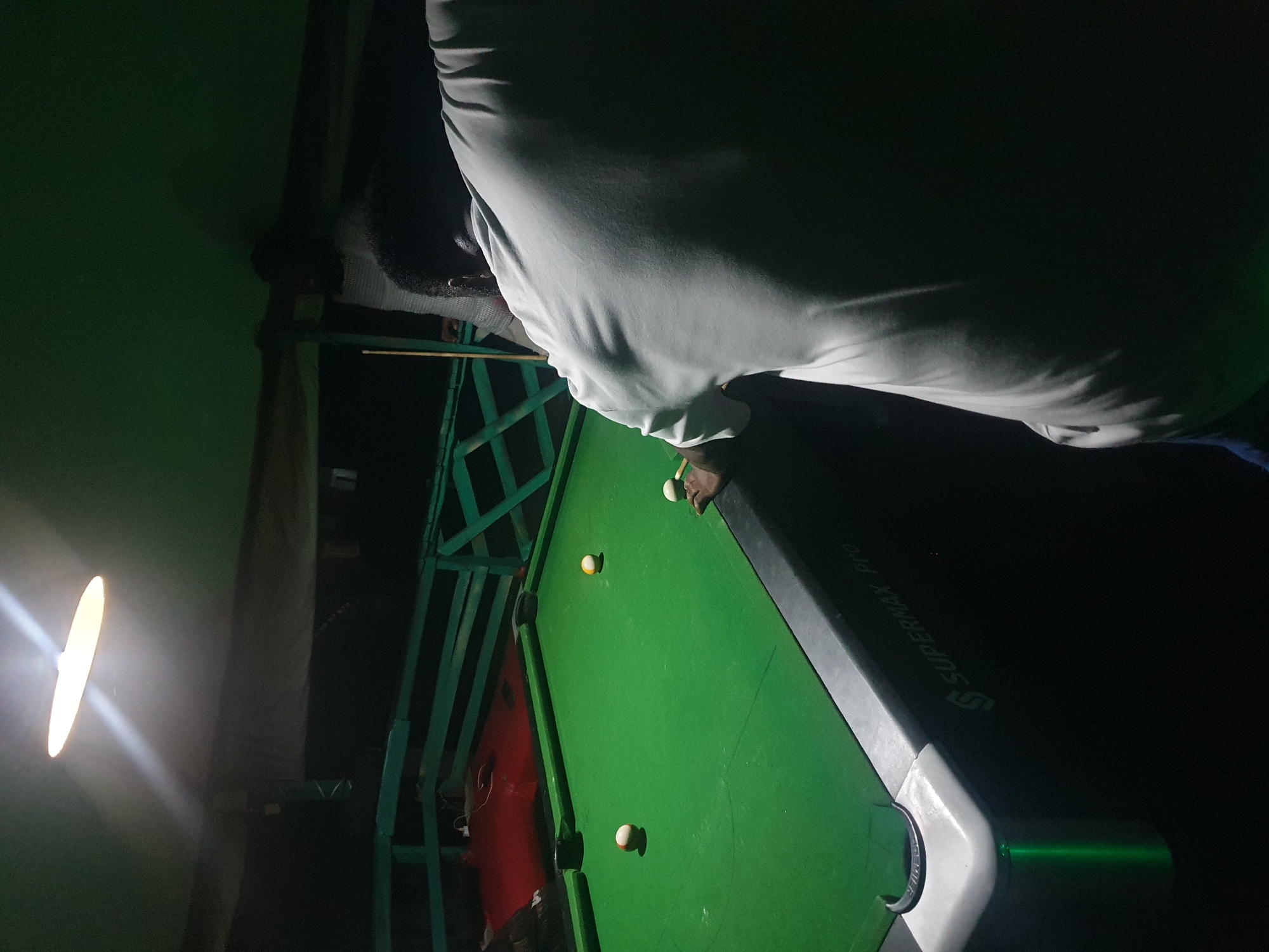 The image depicts a dimly lit room, likely a pool hall in Lokoja, Nigeria, with a green pool table as the central focus. A person, partially visible from the back and right side of the frame, is playing pool. They are wearing a white t-shirt and appear to be in the process of taking a shot, holding a cue stick. Three pool balls are visible on the table: one striped yellow and white ball, one solid orange ball, and another solid white ball. The pool table is a "SUPERMAX Pro" model. In the background, there is a green wooden fence or railing and a red mat or carpeted area. A bright, oval-shaped light fixture hangs from the ceiling, casting illumination and shadows throughout the scene. The overall ambiance is dark and atmospheric, typical of a recreational space at night.