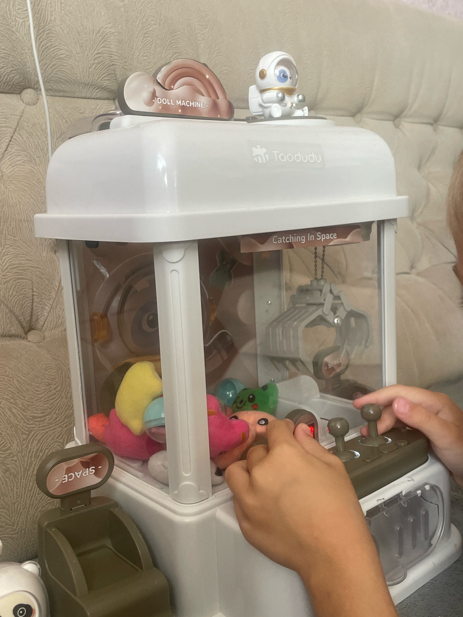 In a domestic indoor setting, likely a living room or playroom, a white claw machine toy, branded "Taodudu," is positioned on a surface. The machine features a transparent compartment filled with small plush toys, including a yellow duck-like toy, a pink toy, and a green frog toy with a smiling face. A child's hands are actively engaged with the machine's controls, manipulating two joysticks. The machine itself is decorated with decals; one reads "- DOLL MACHINE -" and another, featuring a small astronaut figurine, states "Catching in Space." To the left of the machine, a small, dark green toy dispenser with a label reading "- SPACE -" is visible. The background consists of a textured upholstered surface, possibly a sofa or headboard, in a neutral beige color. No specific details pertaining to the city of Baranovka, Russia, are present within the image.