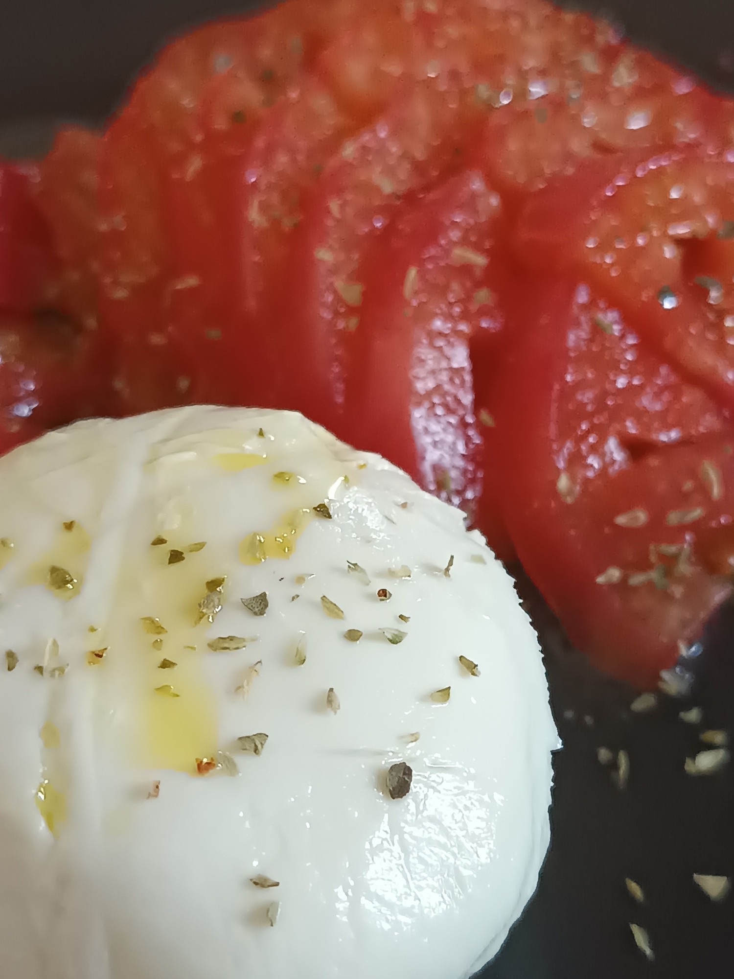 The image shows a close-up of a dish that appears to be caprese salad, consisting of sliced tomatoes and a ball of fresh mozzarella cheese. The tomatoes are a vibrant red and are sliced thinly, fanned out in the background. The mozzarella is a bright white, round ball, generously drizzled with olive oil and sprinkled with dried herbs, possibly oregano or basil. The overall presentation suggests a simple yet appetizing dish. The lighting is somewhat diffused, highlighting the textures of the cheese and tomatoes. There are no people visible in the image. The background is dark and out of focus, emphasizing the food. There is no information in the image that would indicate its location in Viadana, Italy.