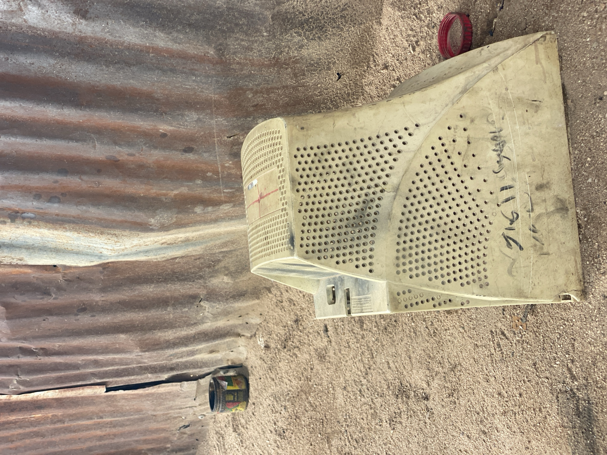 The image depicts an outdoor scene with a corrugated metal wall in the background, appearing weathered and rusted. In the foreground, a dusty, sandy ground is visible. Prominently featured is the beige casing of an old CRT monitor, lying on its side, with its perforated surface facing upwards and outwards. The casing is dirty and shows some signs of wear. To the left of the monitor casing, a small, multi-colored cylindrical object, possibly a can or container, rests on the ground. To the right of the monitor casing, a red circular object, possibly a ring or cap, lies on the sand. There is some writing in black marker on the side of the monitor casing. The overall impression is one of an outdoor storage or refuse area in a place with a sandy environment.