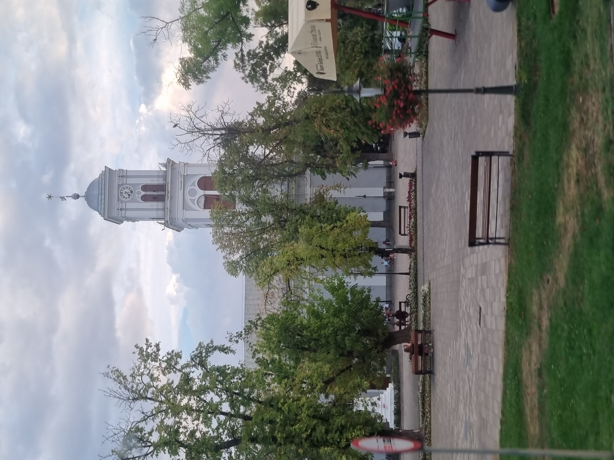 The image depicts a town square in Salonta, Romania, featuring a prominent white church with a clock tower under a cloudy sky. Several trees with green foliage are scattered throughout the square, with some bare branches visible. Paved pathways and grassy areas make up the ground.
In the foreground, a park bench is empty. Further into the square, another bench is occupied by two individuals, one of whom is seated. In the middle ground, a street sign with a red circle and a diagonal line is partially visible on the left. A lamppost with red flowers blooming at its base stands to the right. Behind the trees and church, a white canopy structure with red supports and a playground set are visible on the far right. Cars are parked in the background on the right side of the image. The overall scene suggests a public gathering space in a town.