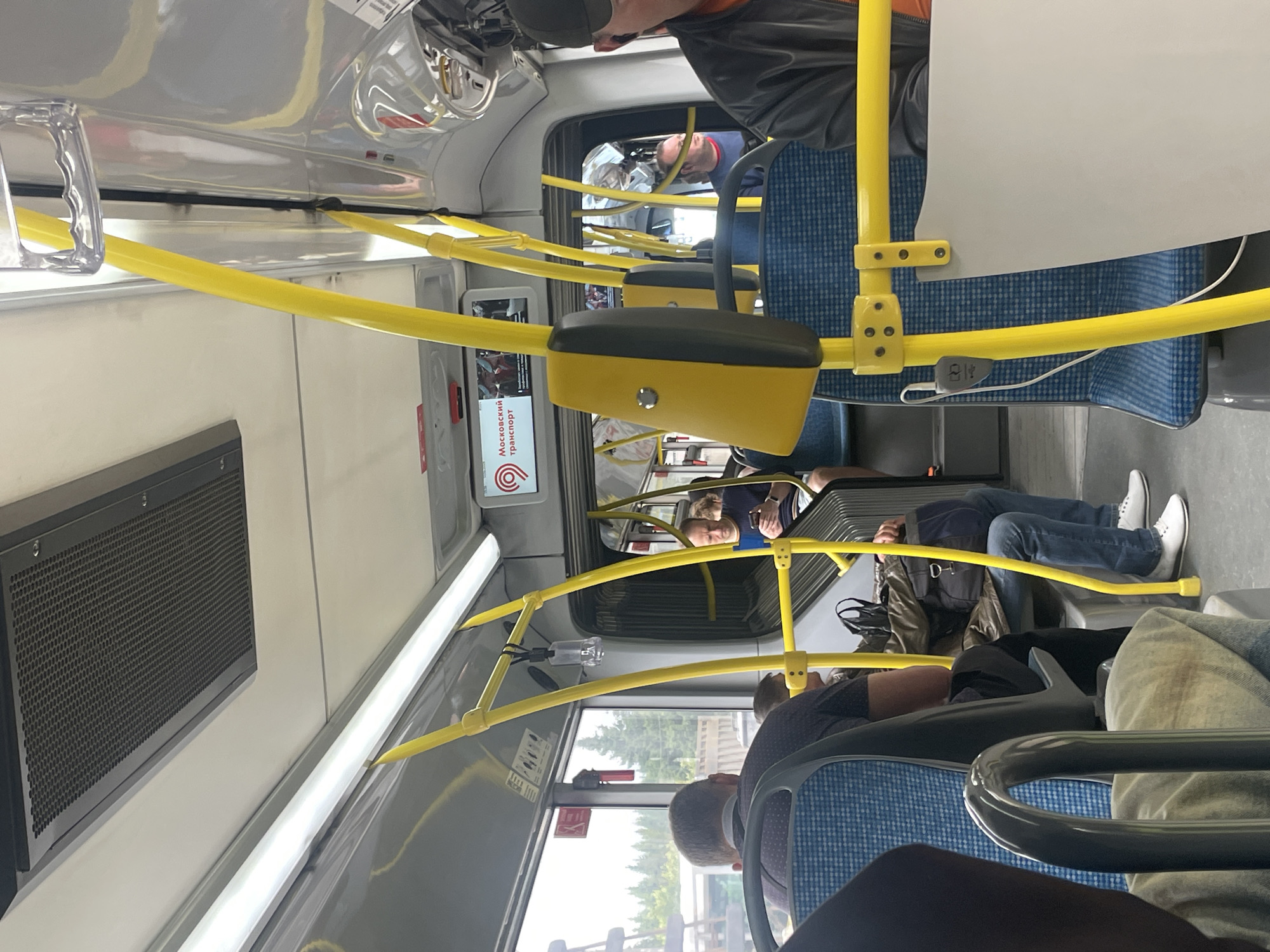 The image shows the interior of a bus in Yakovlevskoye, Russia. The scene is viewed from the perspective of a seated passenger, looking towards the front of the bus. Several people are visible, seated and standing. The bus has blue patterned seats and bright yellow handrails, characteristic of public transportation.
One passenger in the foreground is facing away from the camera, their head and shoulders visible. Further back, a man in a blue shirt and jeans is seated, his legs extended. Another person, possibly also a man, is leaning his head against the window. In the distance, other passengers are partially visible.
A screen on the bus displays text in Cyrillic, which appears to be an advertisement or information related to "Московский транспорт" (Moscow Transport). The lighting suggests it is daytime, and the view through the windows shows trees and a fence, indicating an outdoor environment. The overall atmosphere seems ordinary, depicting a typical bus journey. There are no overt signs of any particular event or strong emotions. Objects like a black vent on the wall and various poles and fixtures of the bus are also visible.