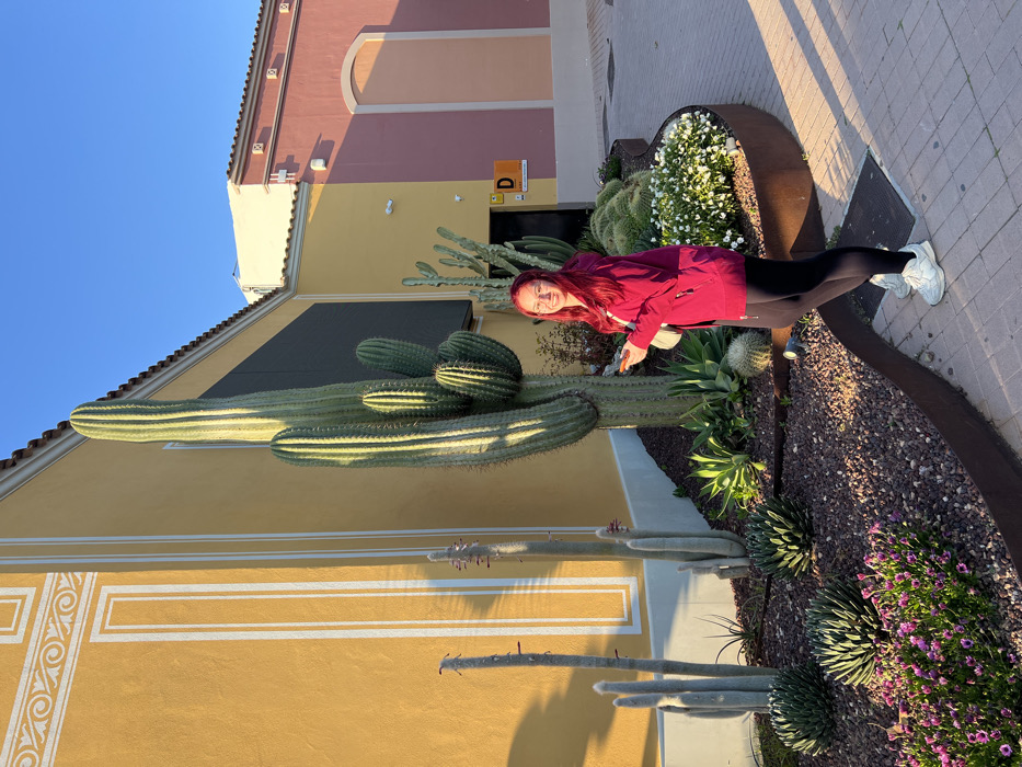 An outdoor scene in Marratxí, Spain, depicts a young adult female positioned centrally, smiling towards the viewer. She has reddish-pink hair, wears glasses, a magenta hooded jacket, black leggings, and white athletic shoes. A light-colored smartphone is held in her right hand, and a white shoulder bag is worn across her body. She stands on a light-colored paver walkway, adjacent to a raised garden bed filled with dark gravel and edged with rusted metal. The garden features a prominent, tall, multi-branched columnar cactus, several smaller columnar cacti including fuzzy white varieties, various succulents such as agave and round cacti, and flowering groundcover with purple and white blooms. Behind the garden is a bright yellow building with white architectural framing and scrollwork, along with a dark rectangular screen. To the right, another building section displays pale yellow and dusty rose facades, featuring an arched recess and a visible 'D' sign. The clear blue sky and prominent shadows indicate direct sunlight.